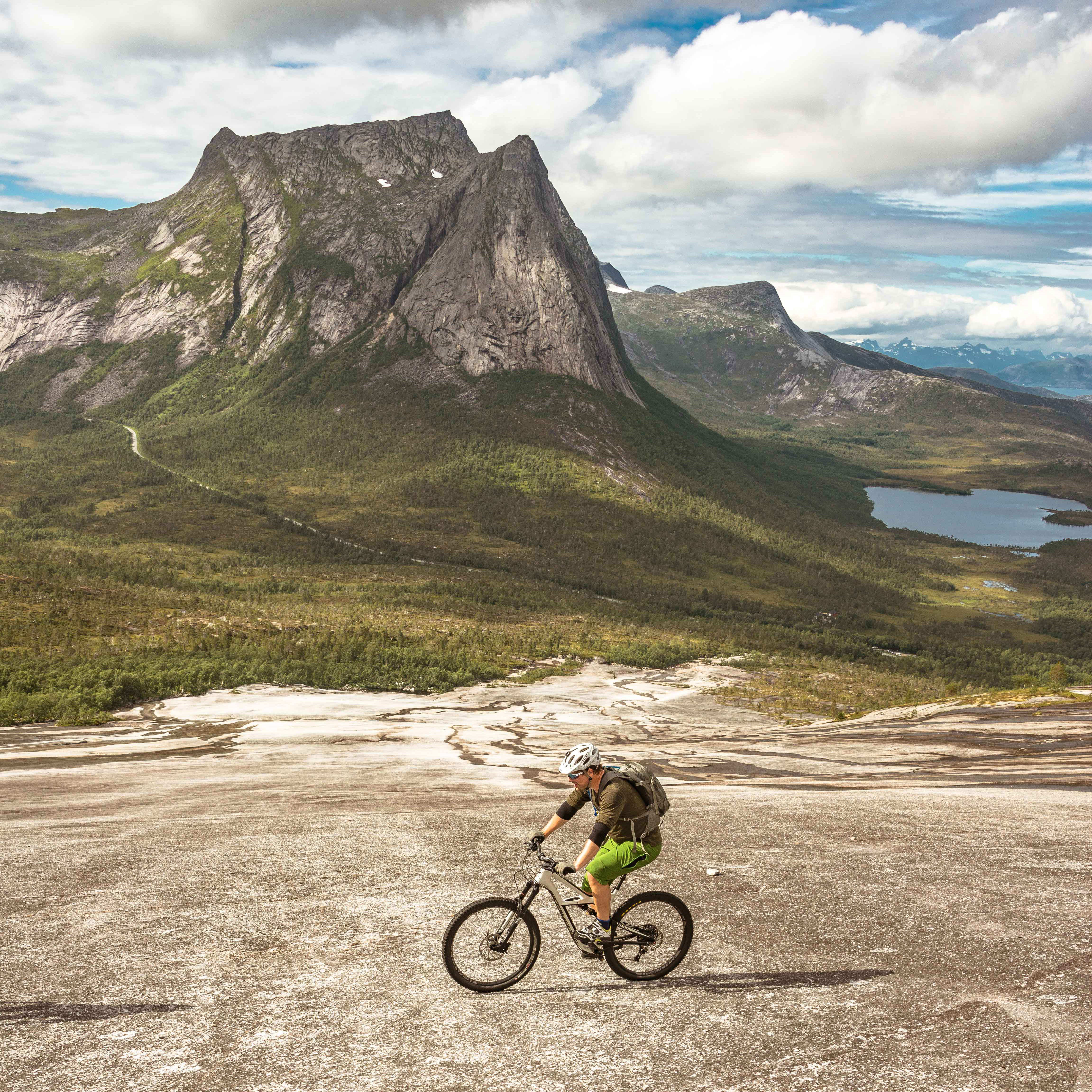 A person wearing Norrøna clothing is riding a mountain bike down a paved road. The road is in the foreground with a mountainous background.