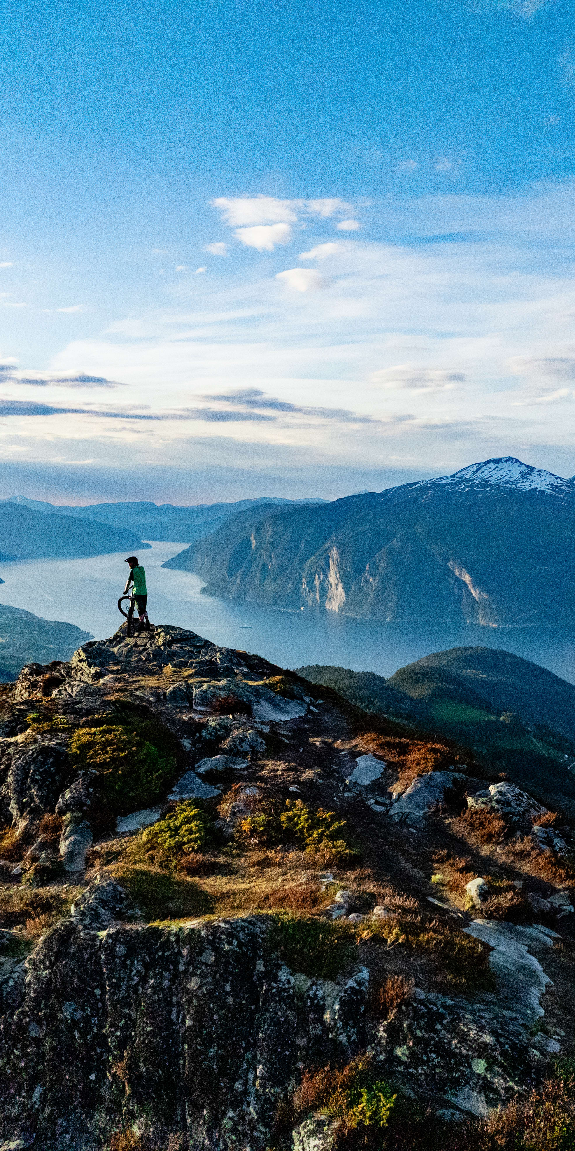 A mountain biker in Norrøna gear stands on a rocky peak overlooking a scenic fjord.