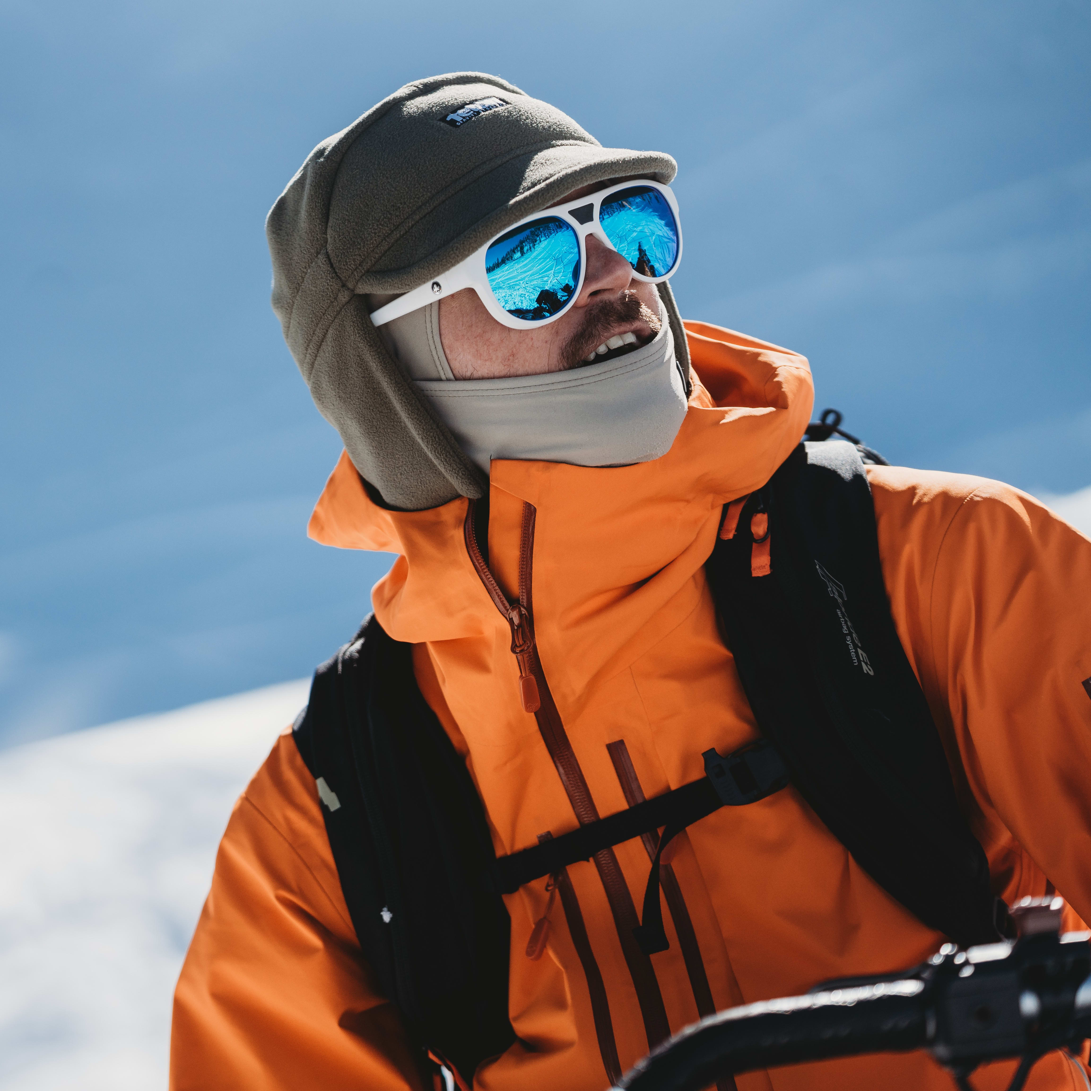 Person wearing an orange Norrøna jacket, winter hat, and blue reflective sunglasses against a clear sky.
