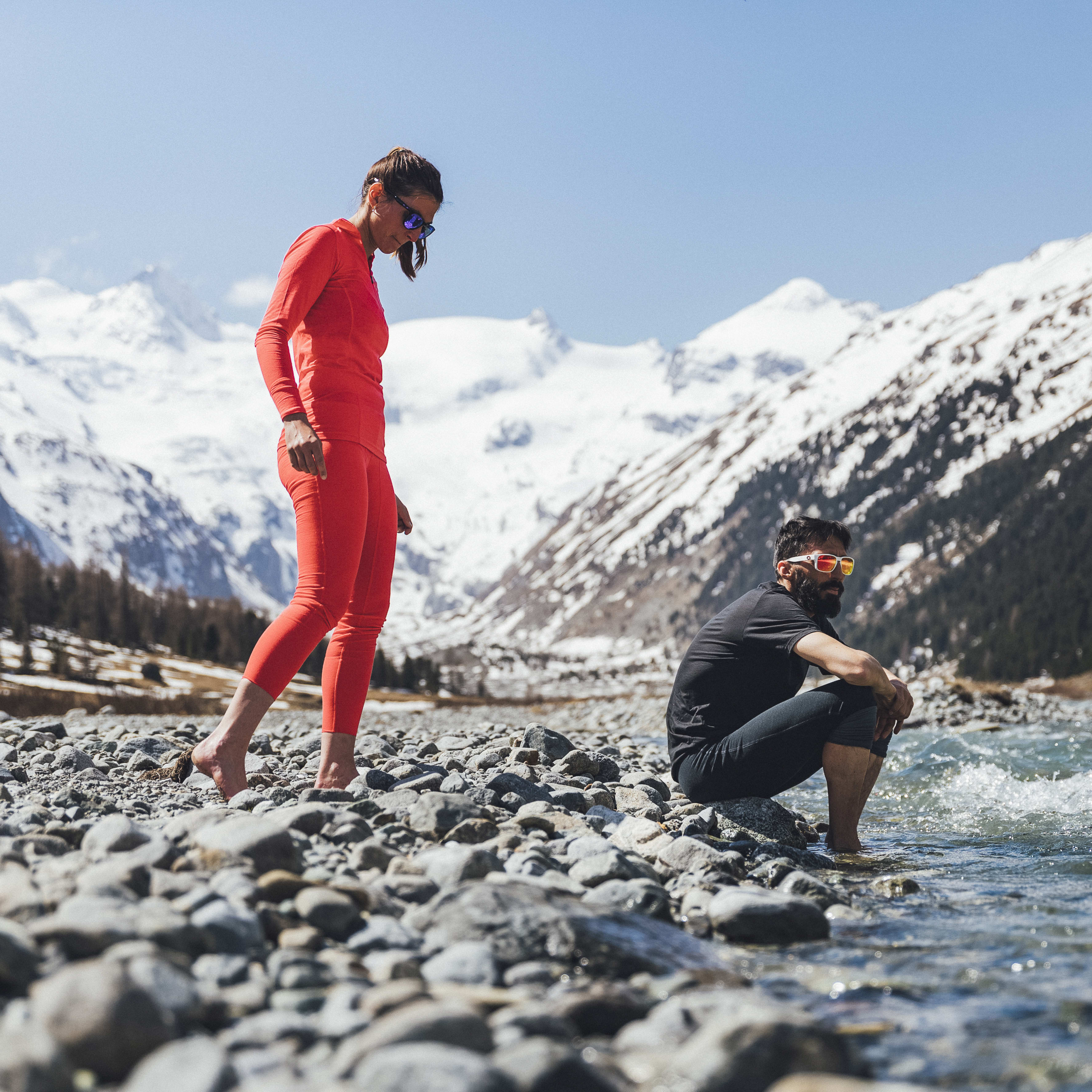 Two people near a river with snowy mountains behind them. One wears a red Norrøna base layer set, the other sits barefoot in the water.
