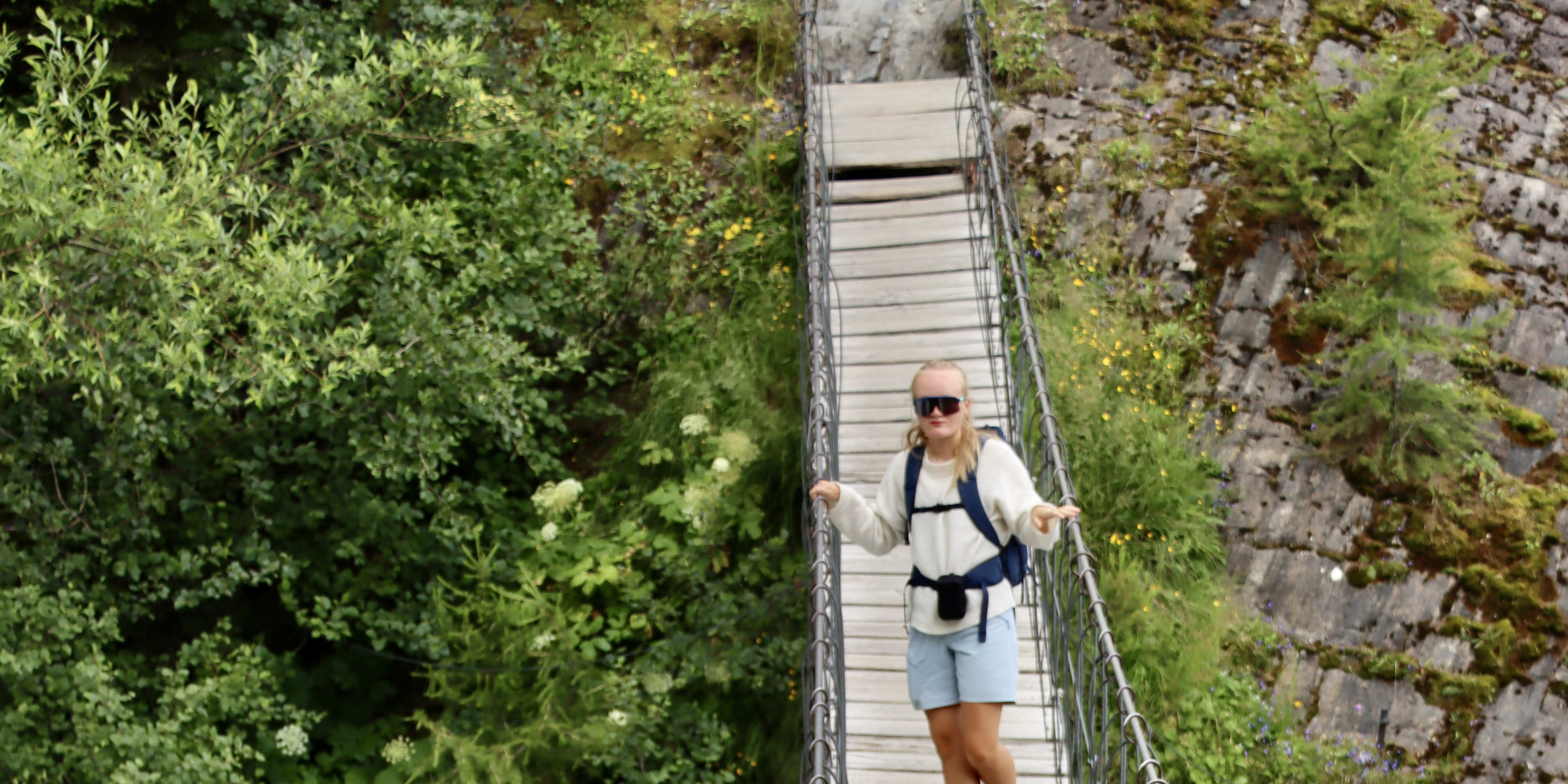 A woman walks on a suspension bridge, wearing sunglasses, a white sweater, a Norrøna backpack and blue shorts with a river beneath.