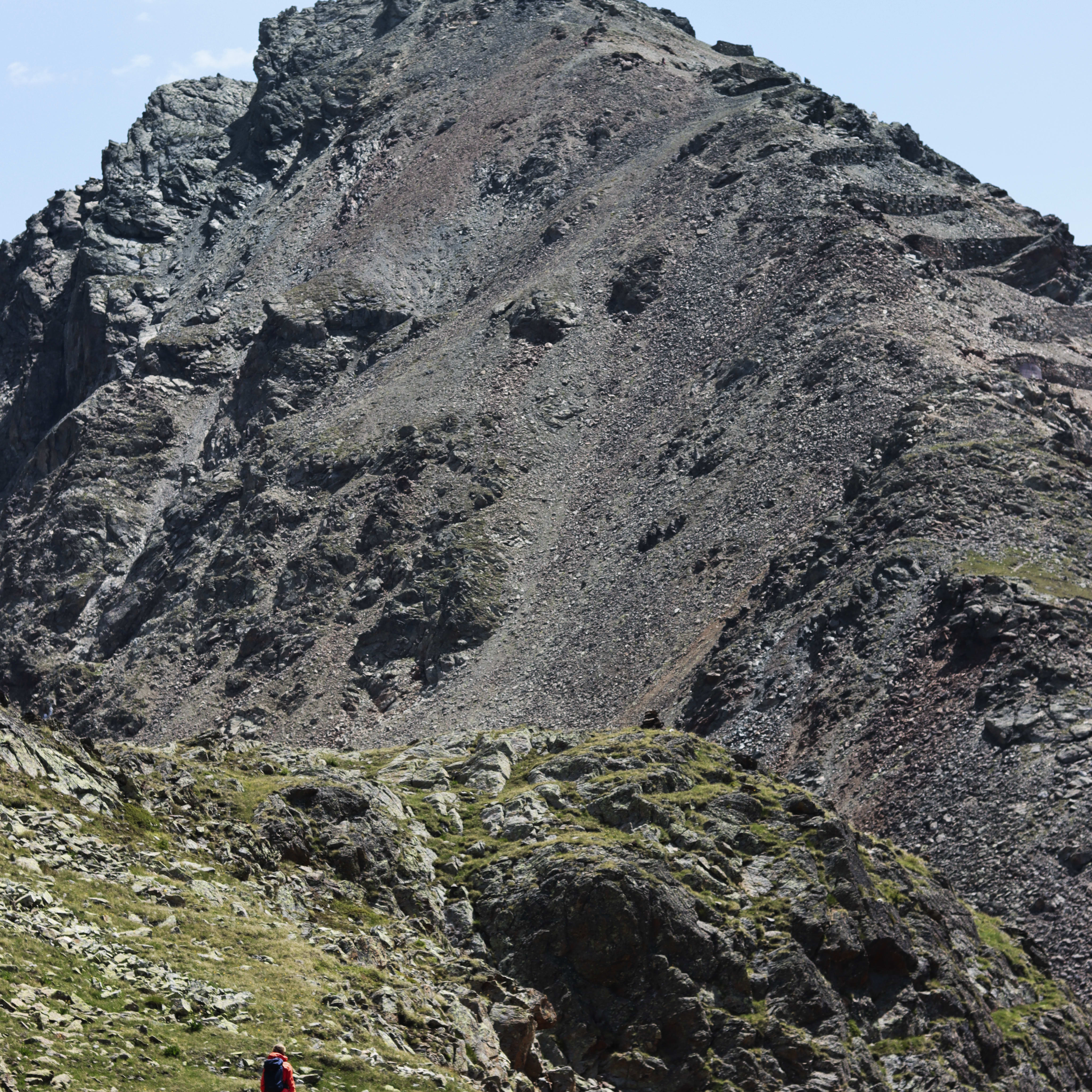 A hiker in a Norrøna jacket walks on a mountain trail leading up to a rocky peak.