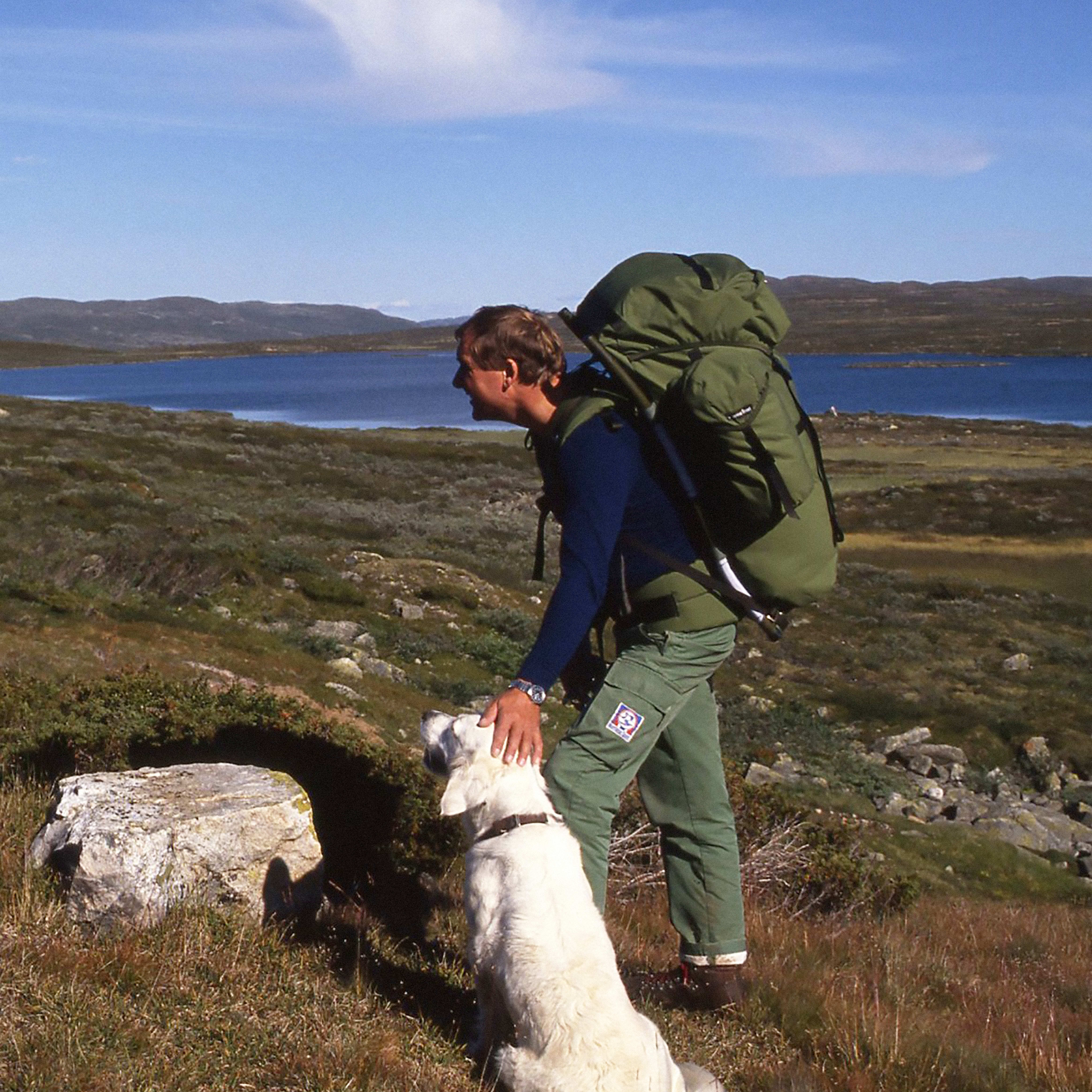 A hiker in Norrøna pants and a large green backpack pets a white dog in a hilly landscape with a lake. Blue sky.