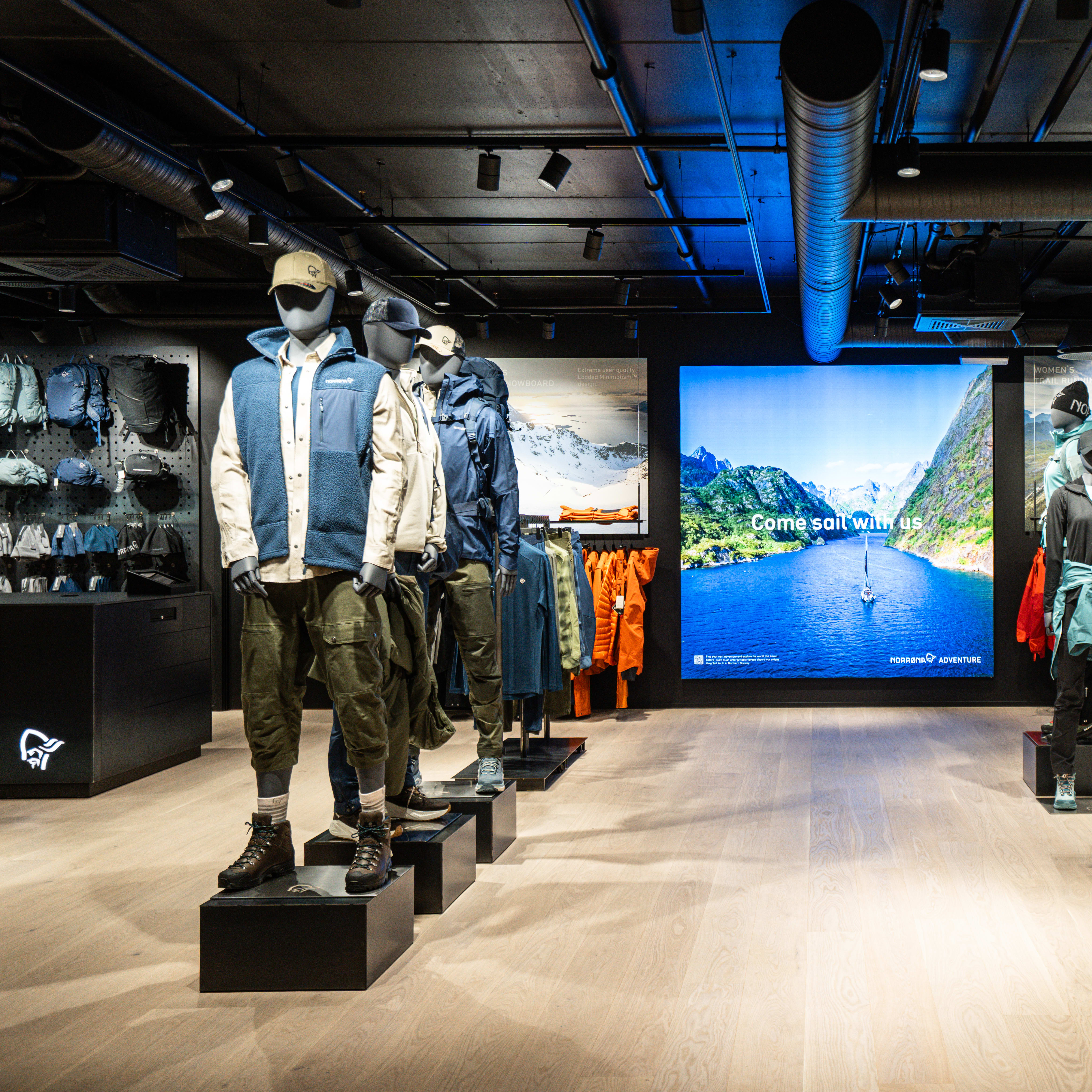 Store interior showcasing Norrøna apparel on mannequins. A large screen displays a fjord view. Clothes racks and folded garments are also visible.