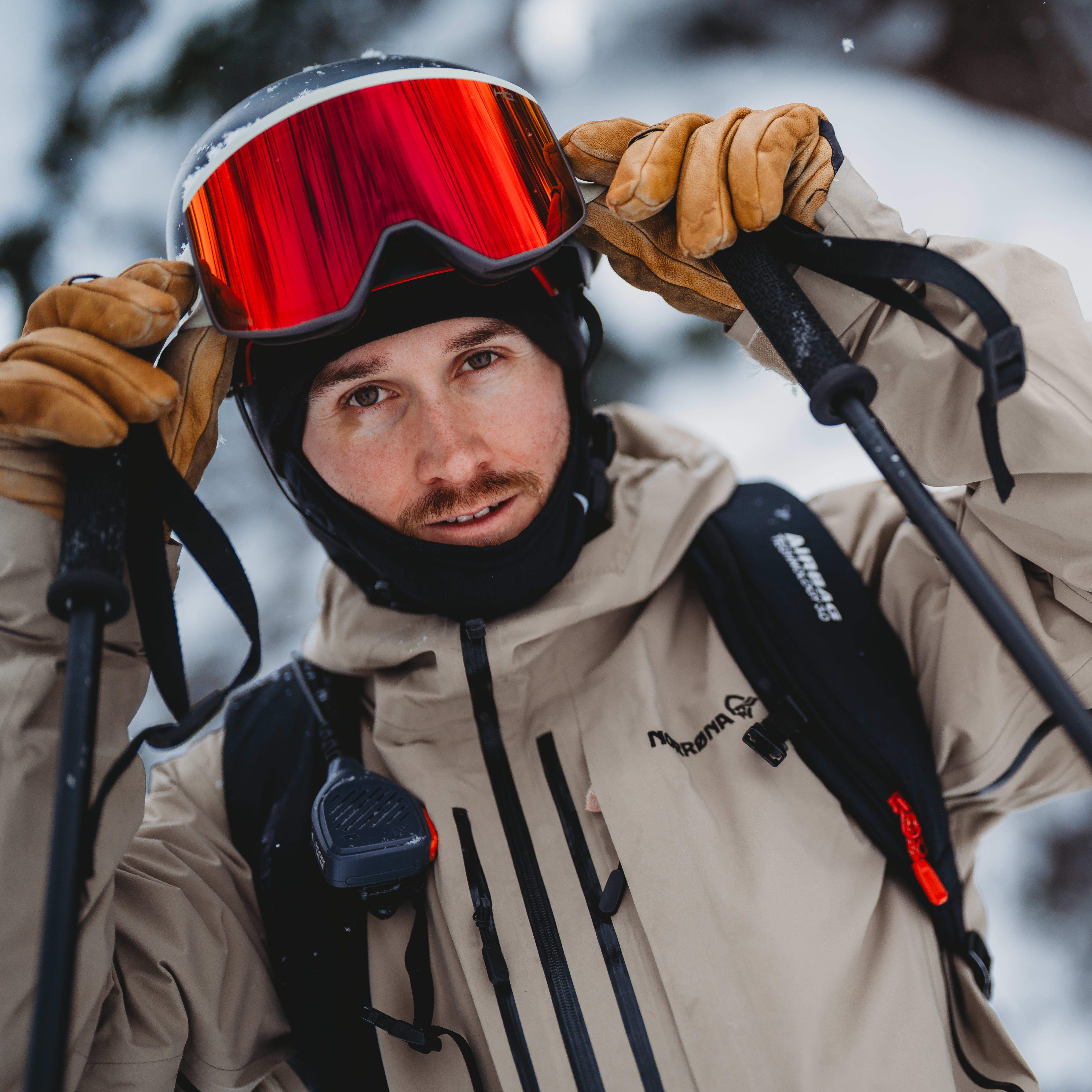Skier wearing a beige Norrøna jacket, helmet, and red goggles, holding ski poles.