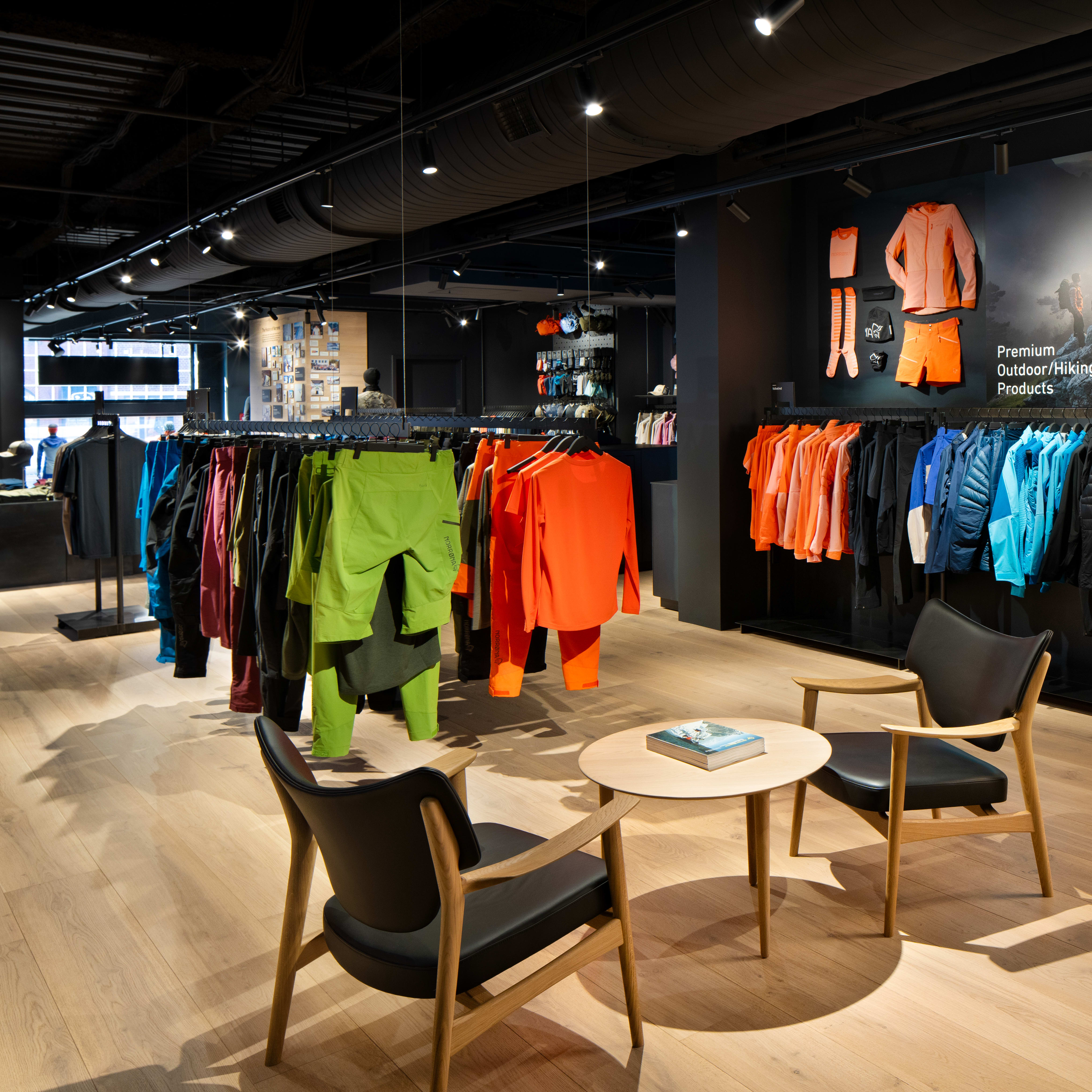 A well-lit Norrøna store with clothing on display, two chairs and a table, with a large poster behind the clothing rack featuring a sunset and mountains in the background. 
