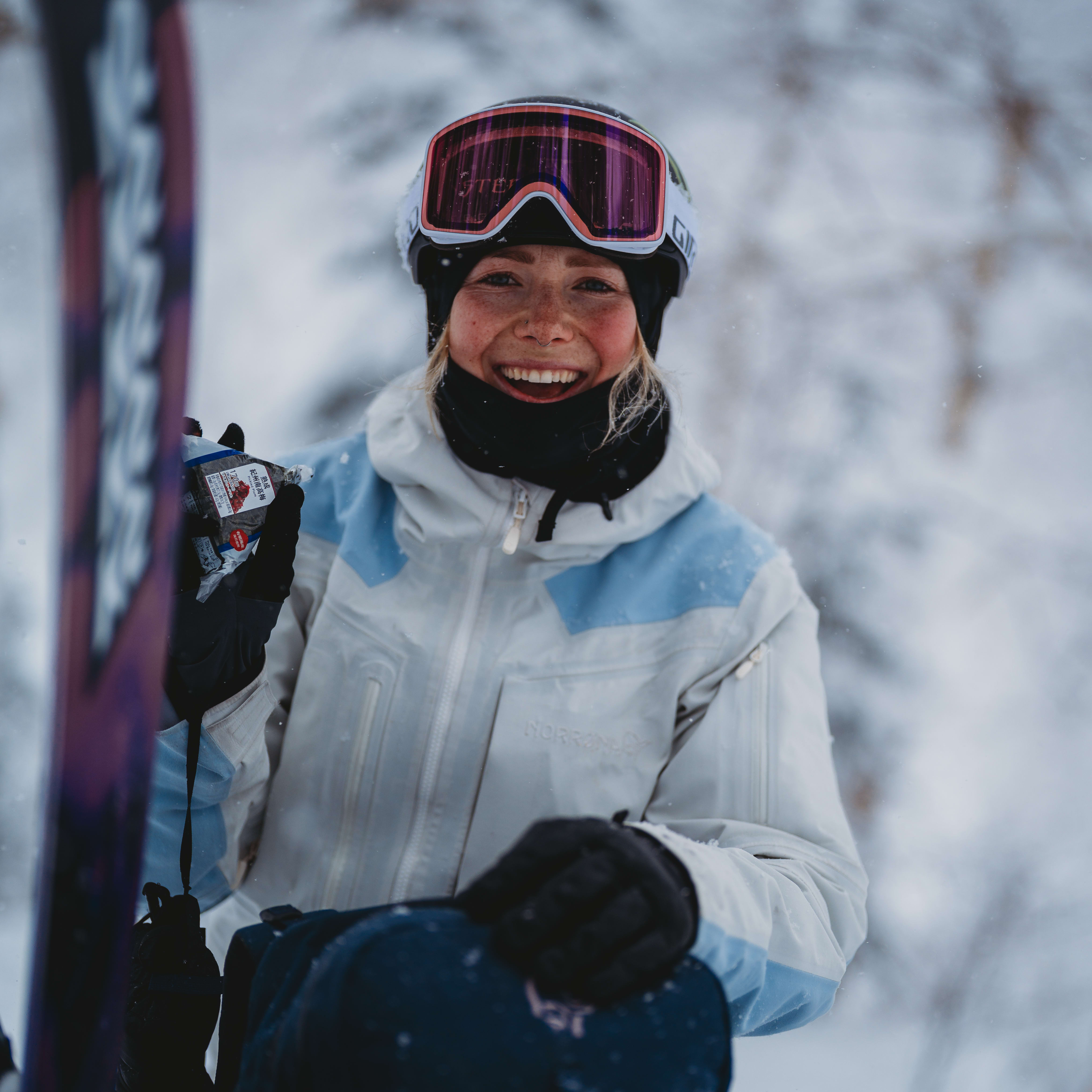 Smiling skier wearing a Norrøna jacket and goggles holds a snack in the snow.