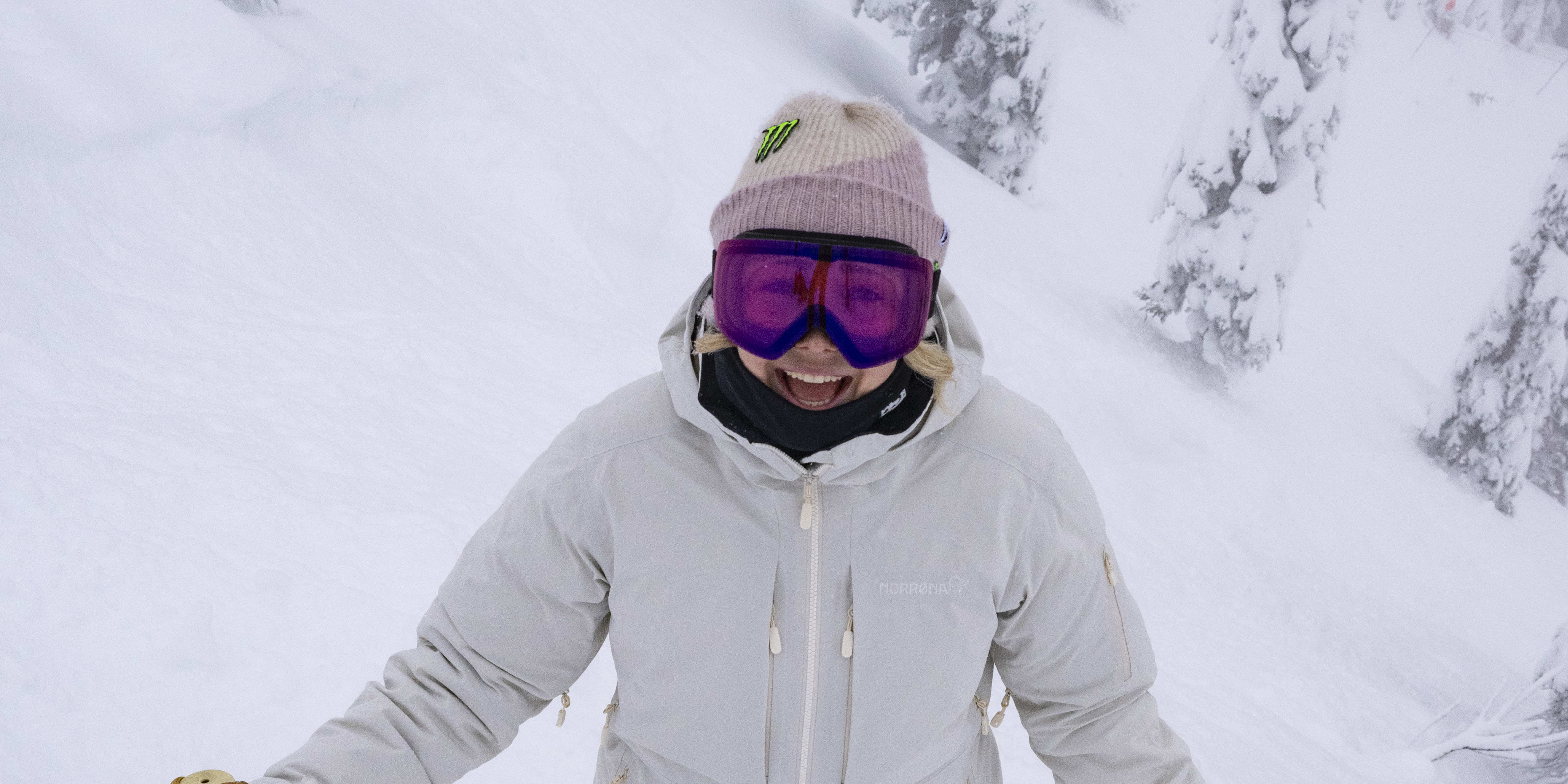A woman wearing a Norrøna jacket and ski goggles smiles while holding ski poles in snowy mountain terrain.