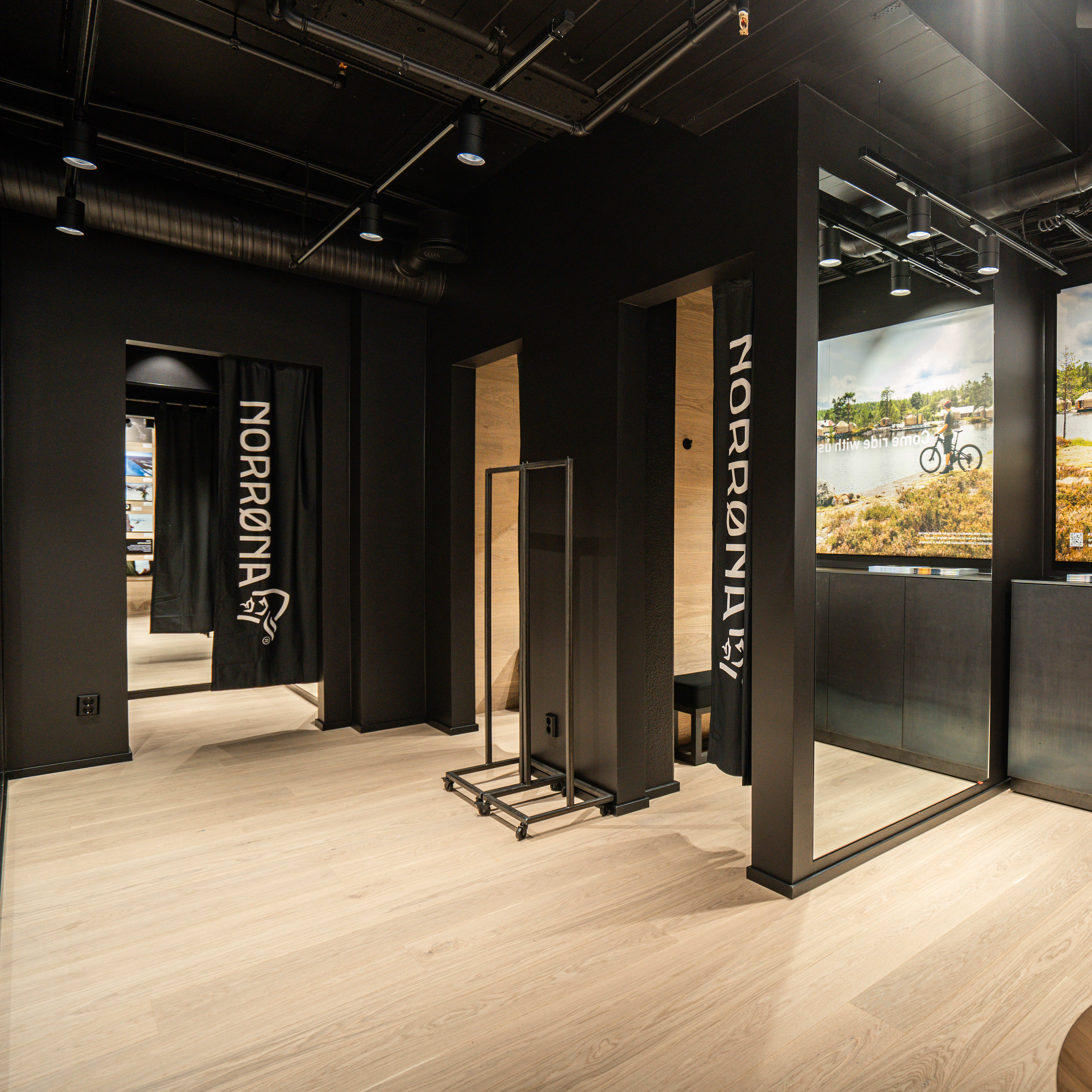 A Norrøna store fitting room area featuring black walls, light wood floors, mirrors, and a nature-themed billboard.