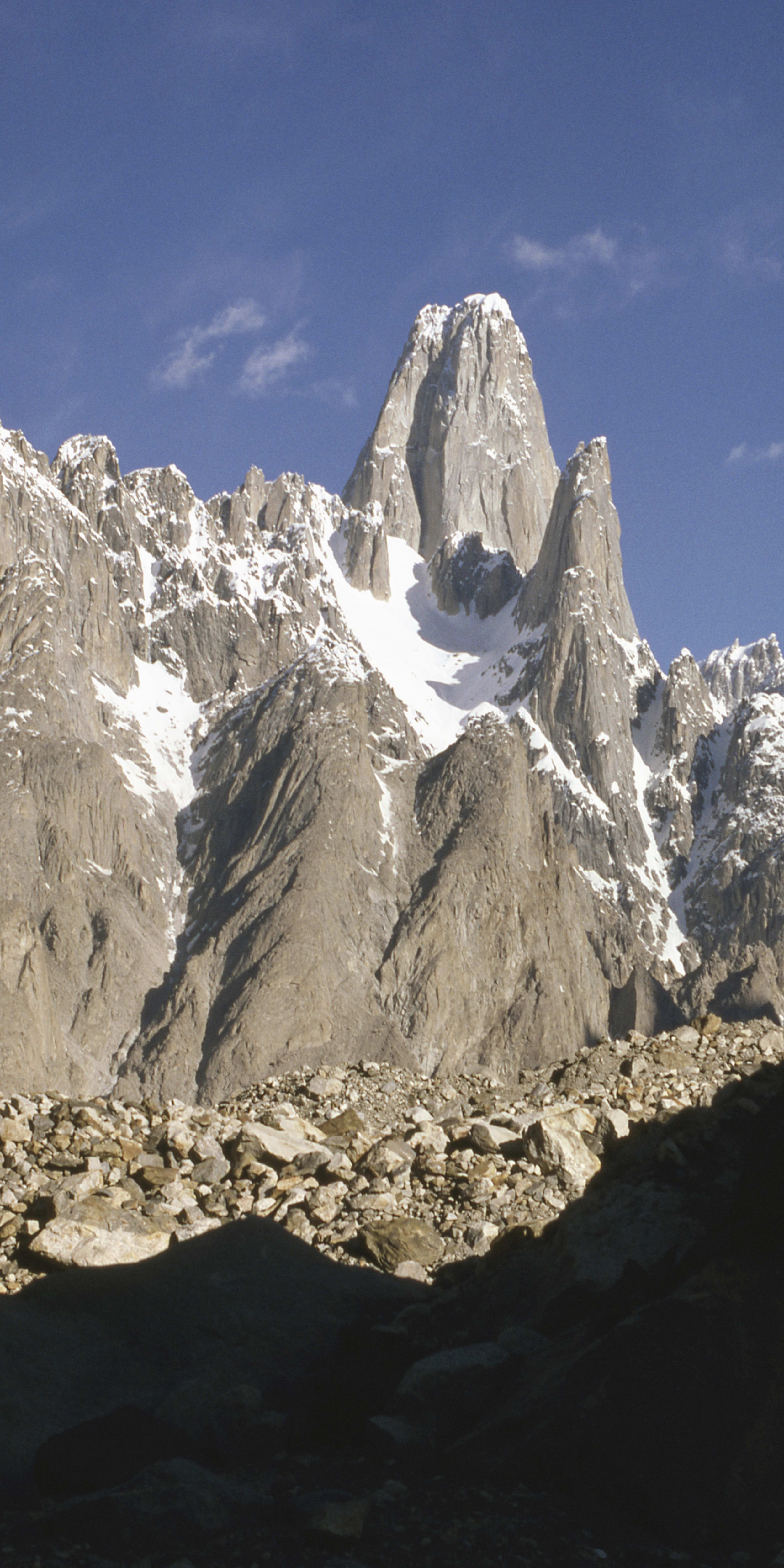 A tall, snow-capped mountain peak rises against a clear blue sky. The mountain is rocky and rugged with patches of snow. The foreground is a rocky, uneven landscape.