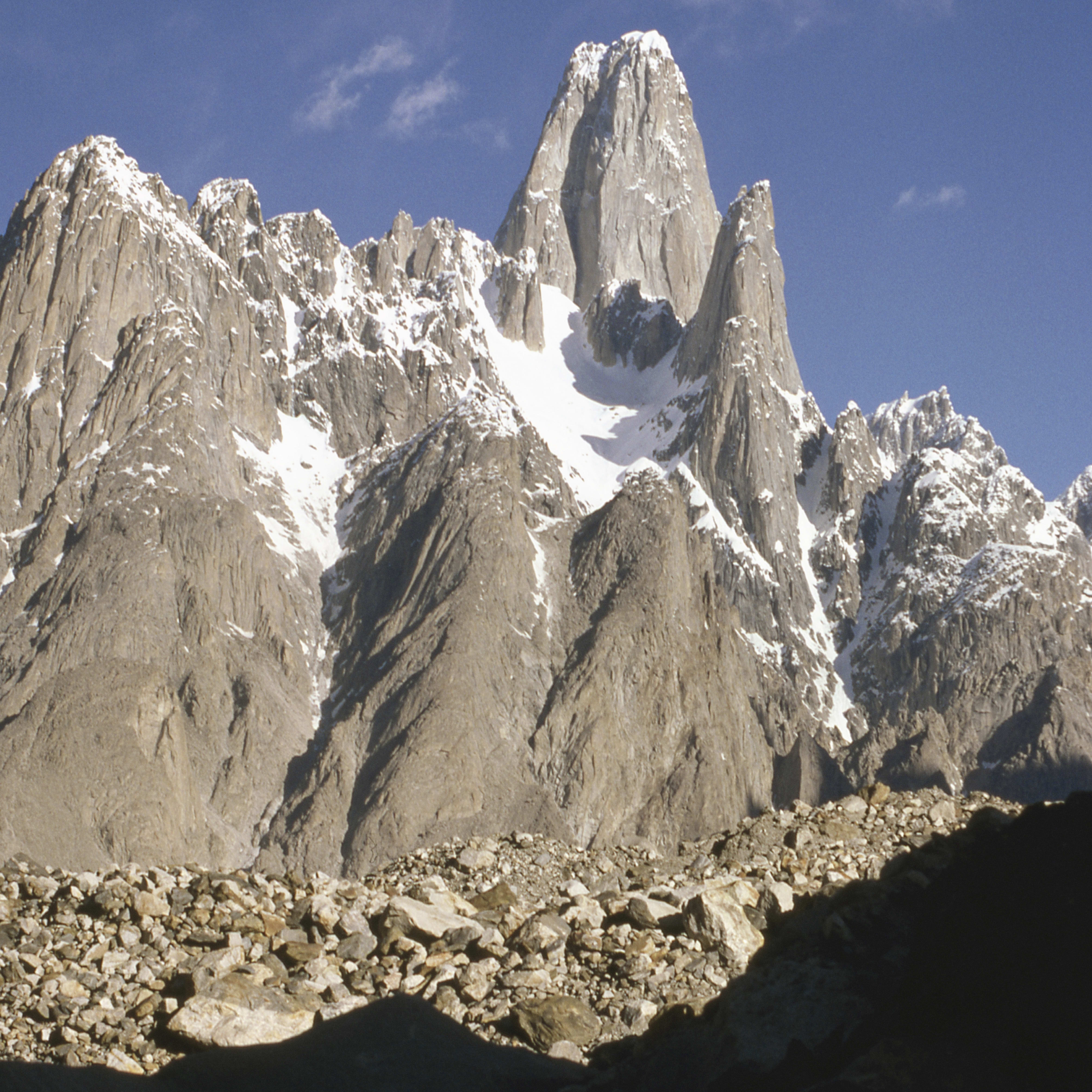 A tall, snow-capped mountain peak rises against a clear blue sky. The mountain is rocky and rugged with patches of snow. The foreground is a rocky, uneven landscape.