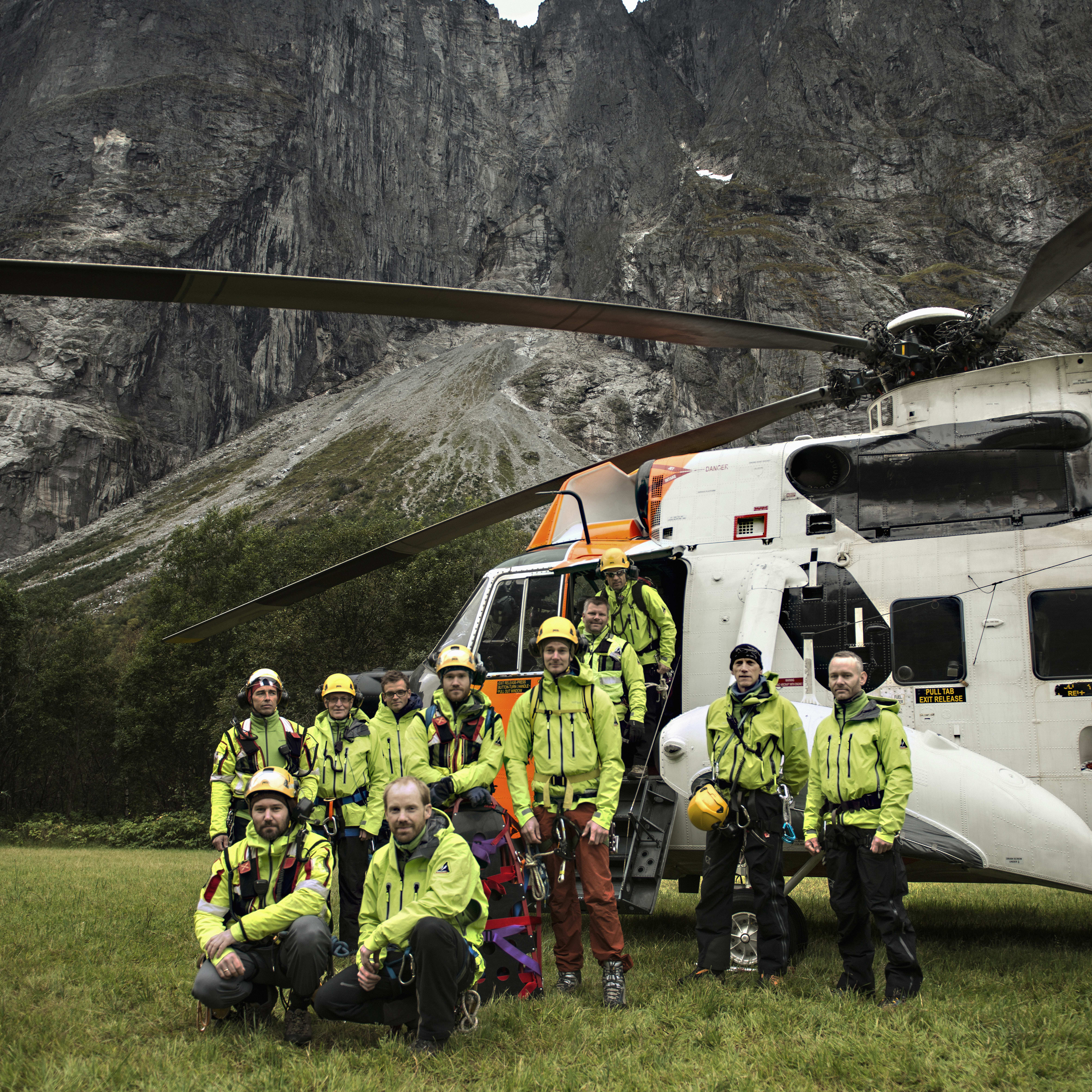 A rescue team stands before a helicopter and towering mountains. The team wears Norrøna jackets and helmets.