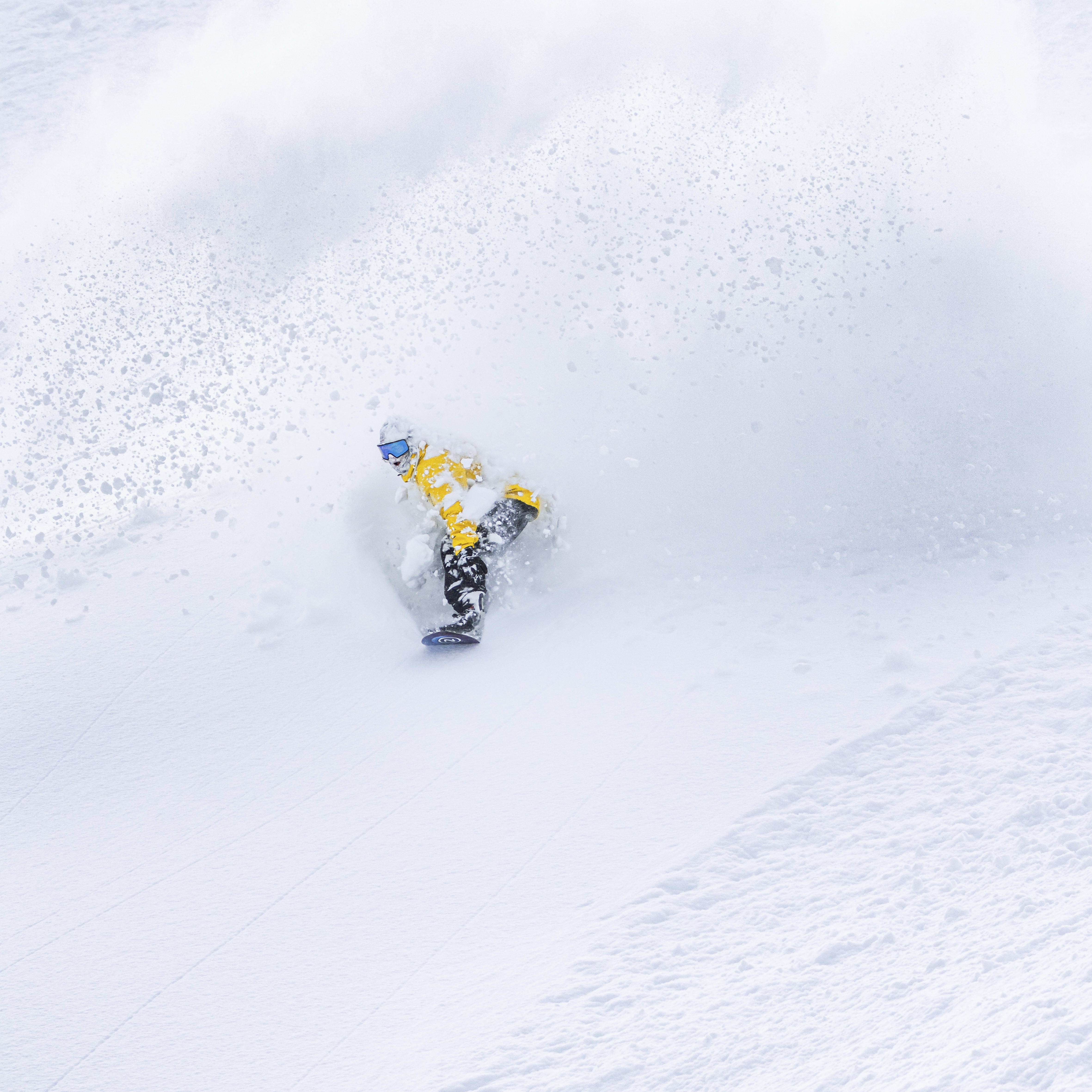 A snowboarder in a yellow jacket kicks up a spray of snow while riding downhill.