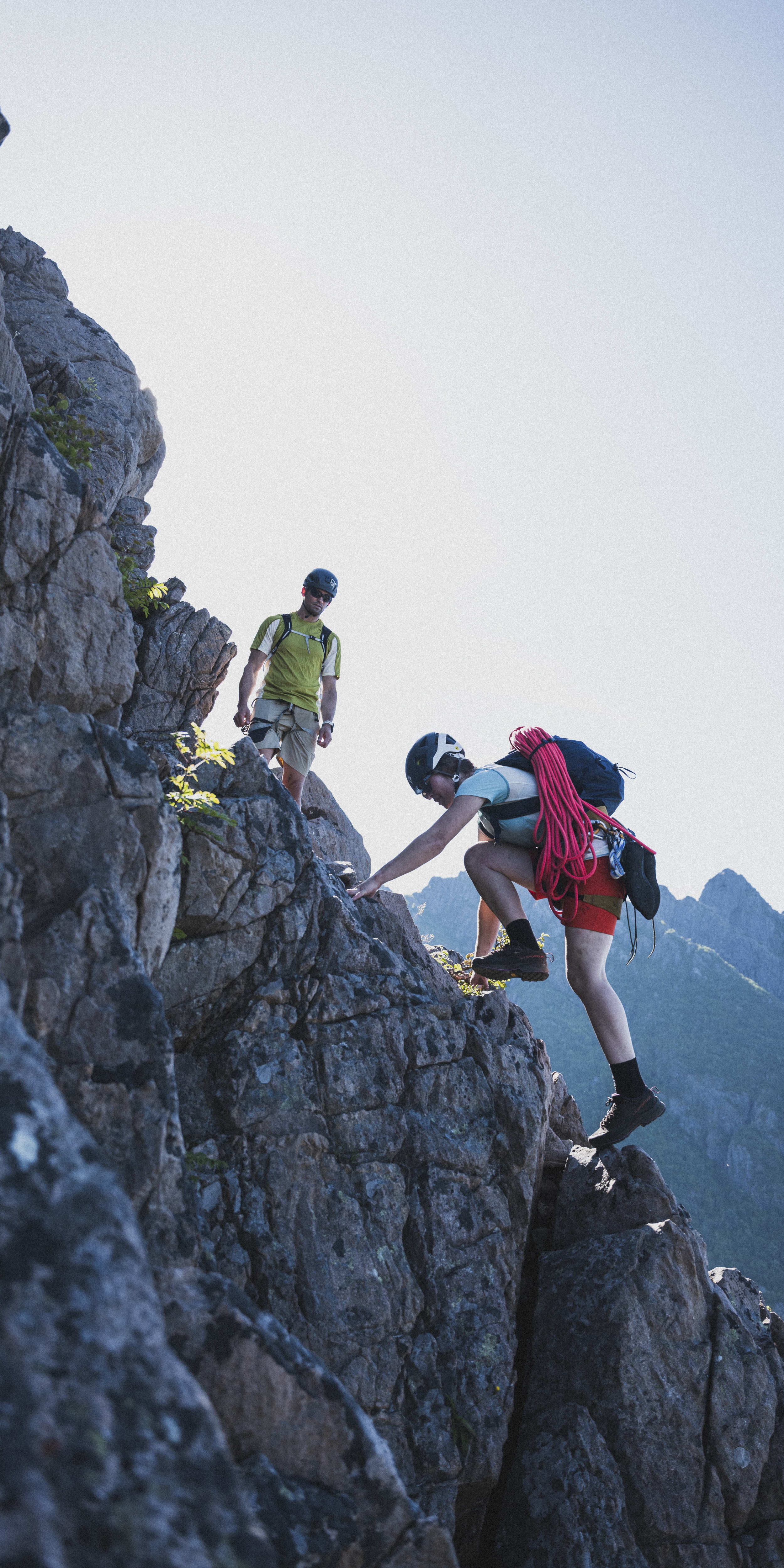 Two climbers navigate a rocky cliff face against a clear sky and mountain backdrop. The lead climber, wearing a helmet, a Norrøna backpack and red shorts, ascends.