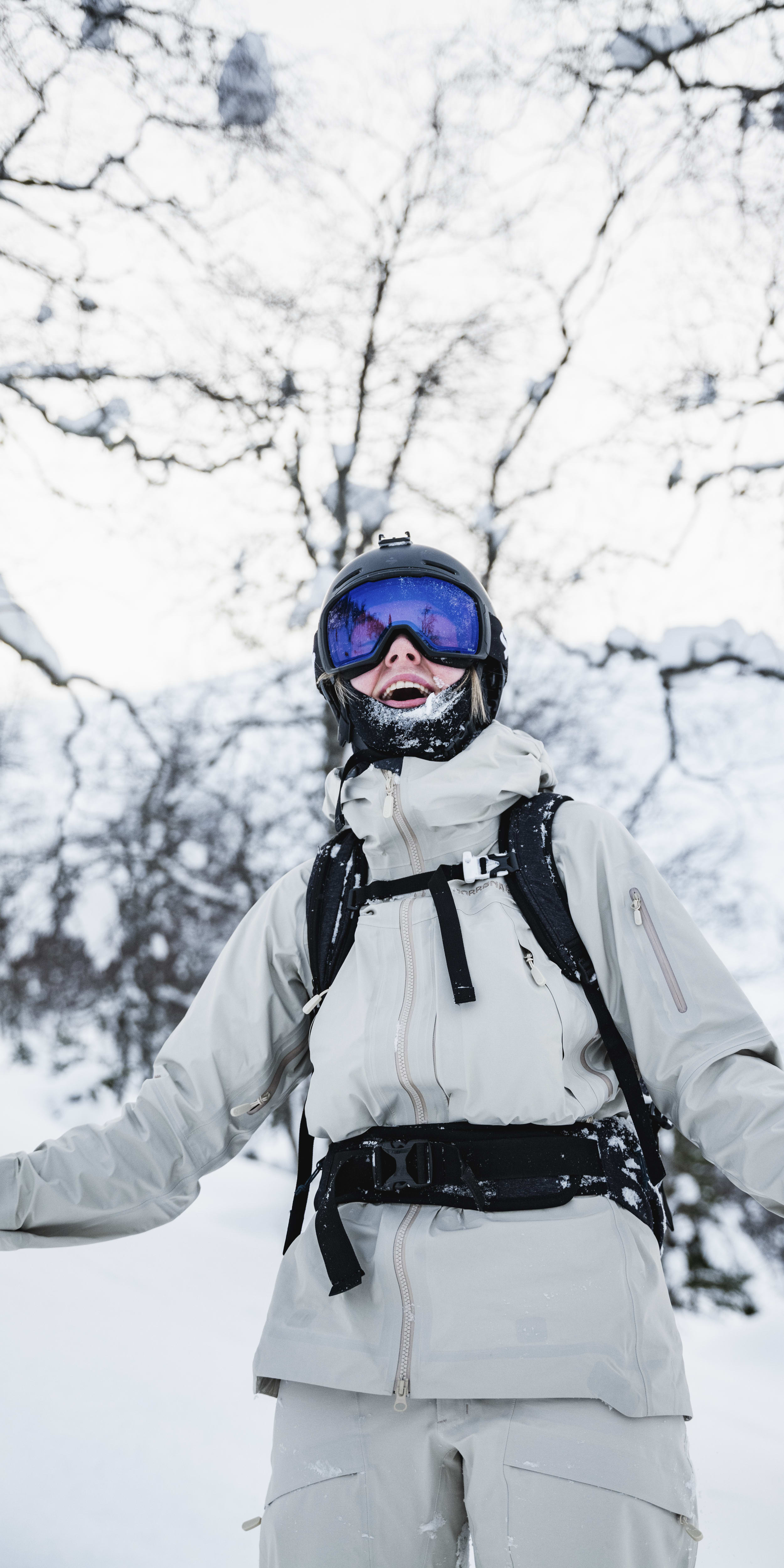 A person wearing a Norrøna ski jacket and pants smiles for the camera in a snowy forest.