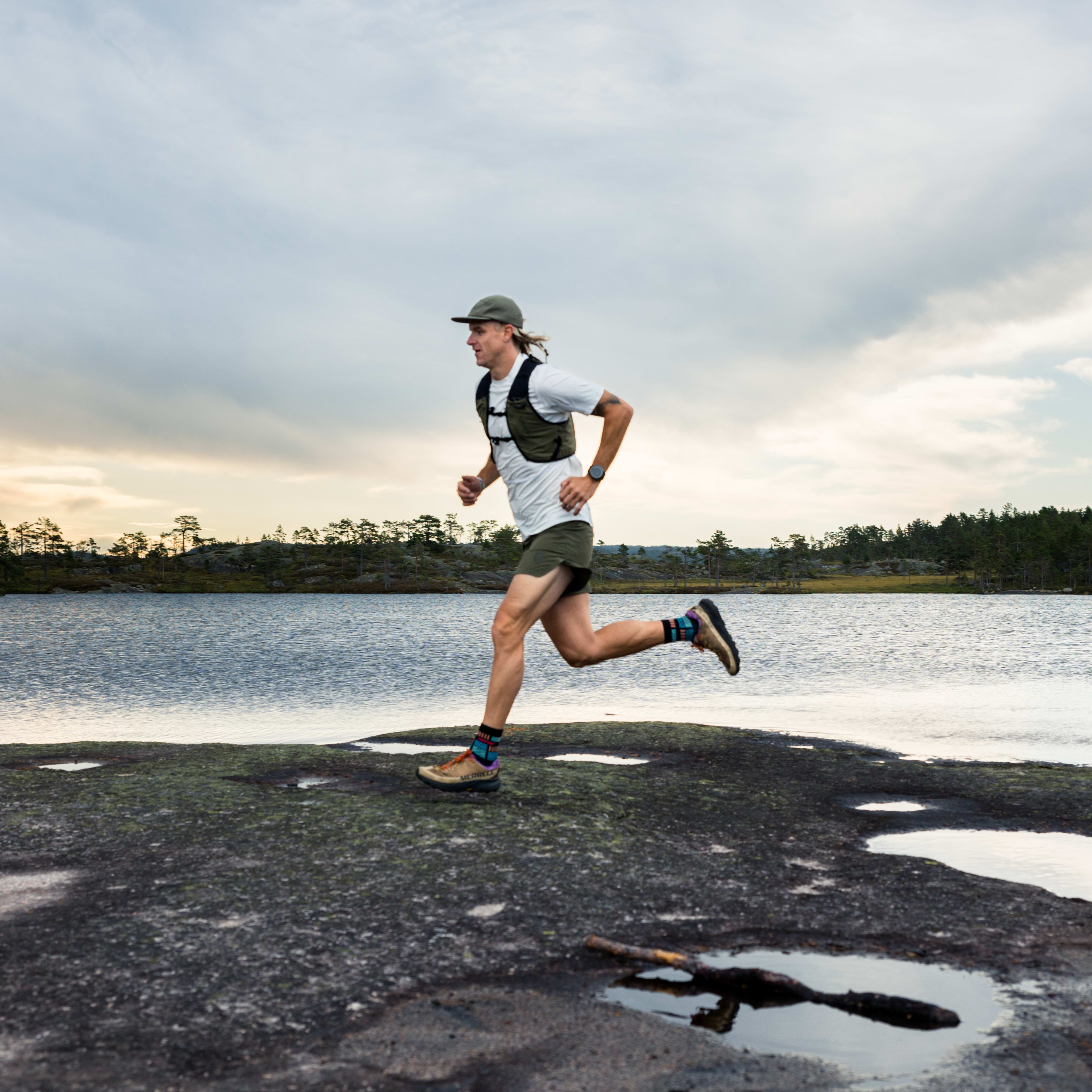 A runner in Norrøna shorts and vest runs near water under a cloudy sky.