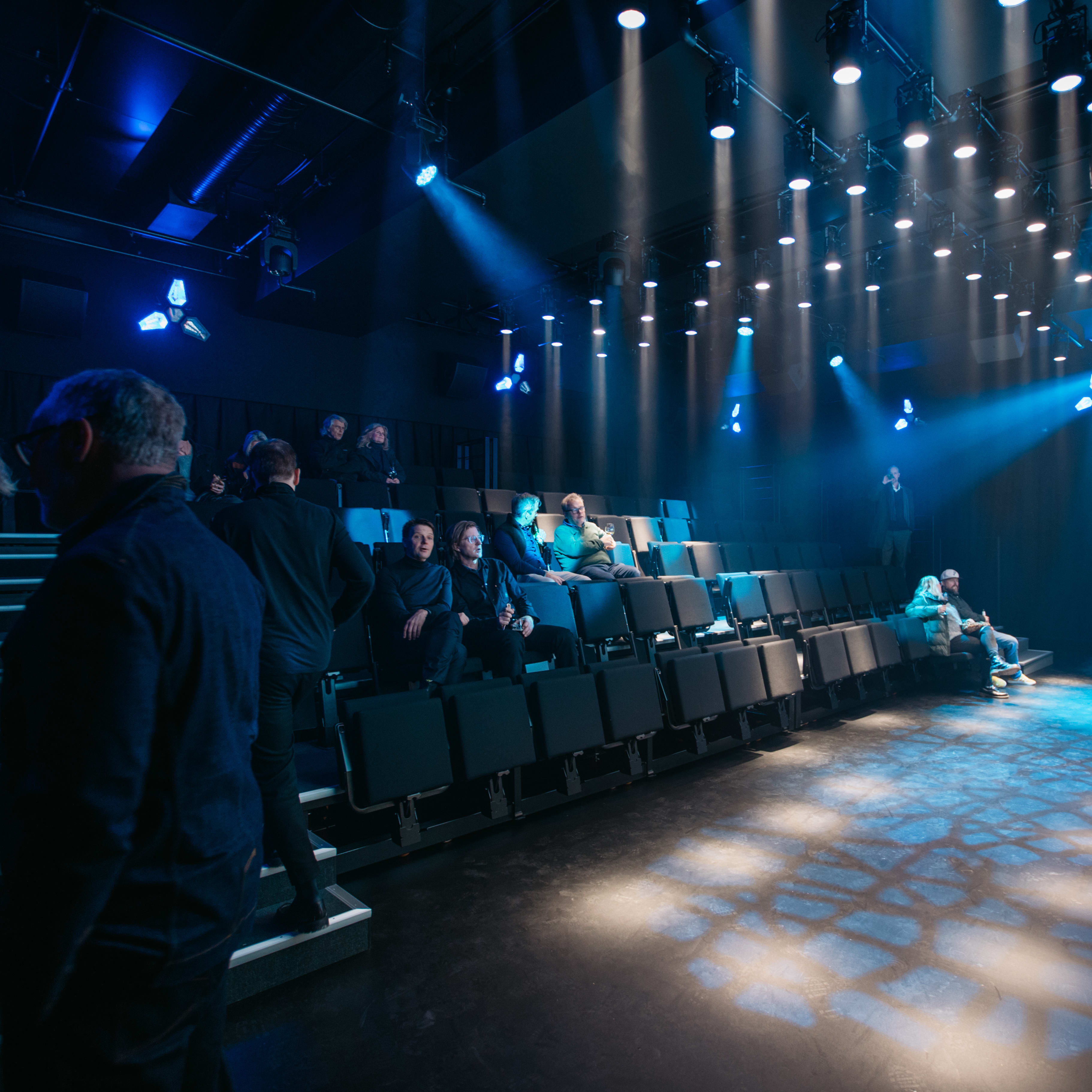 A group of people sitting in a dark theater with blue spotlights shining on them. The people are wearing Norrøna clothing. A large screen is behind them, with more blue lights on the ceiling above.