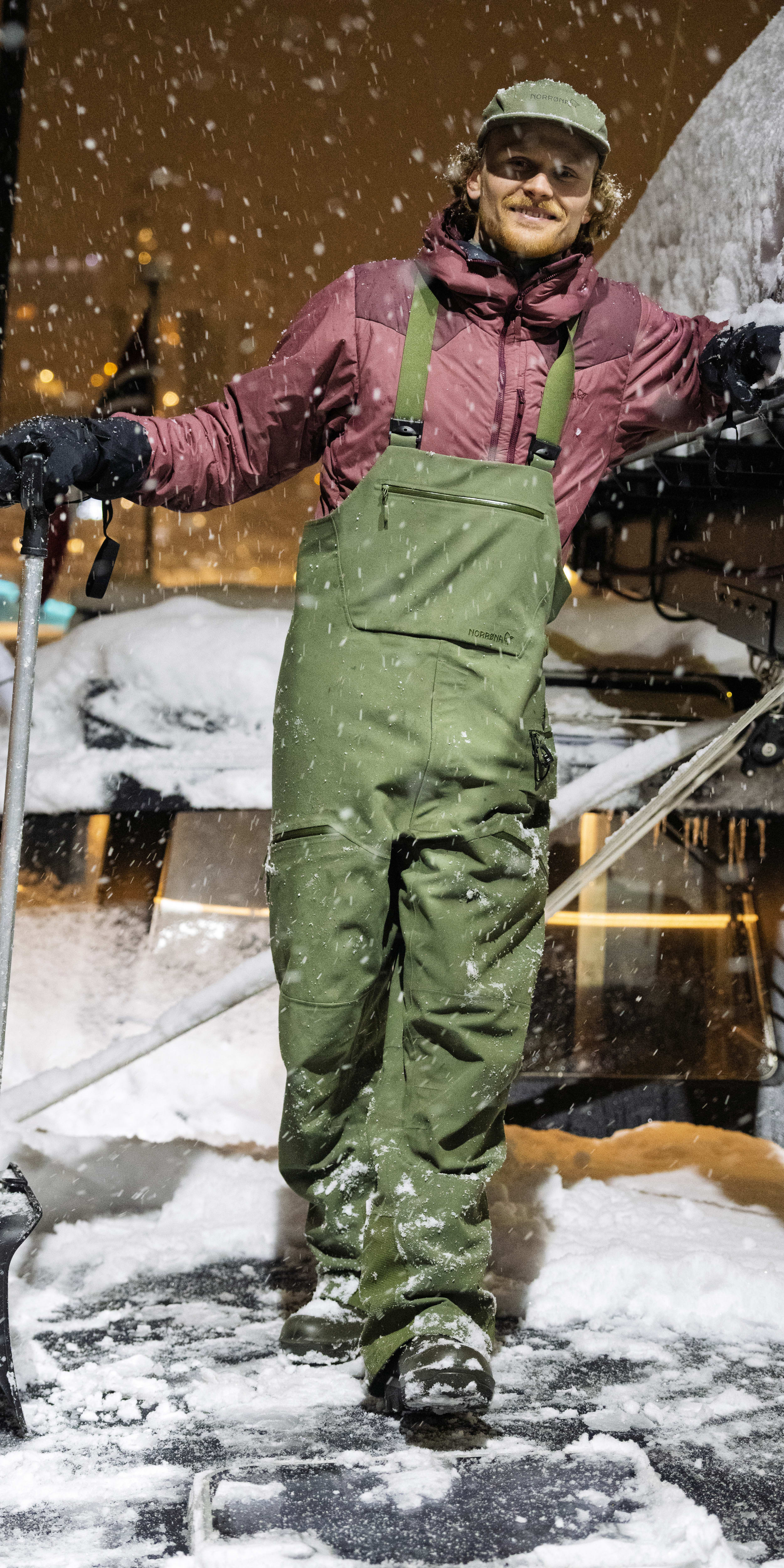 A smiling man shovels snow wearing a Norrøna jacket, hat, and bib overalls on a snowy night.