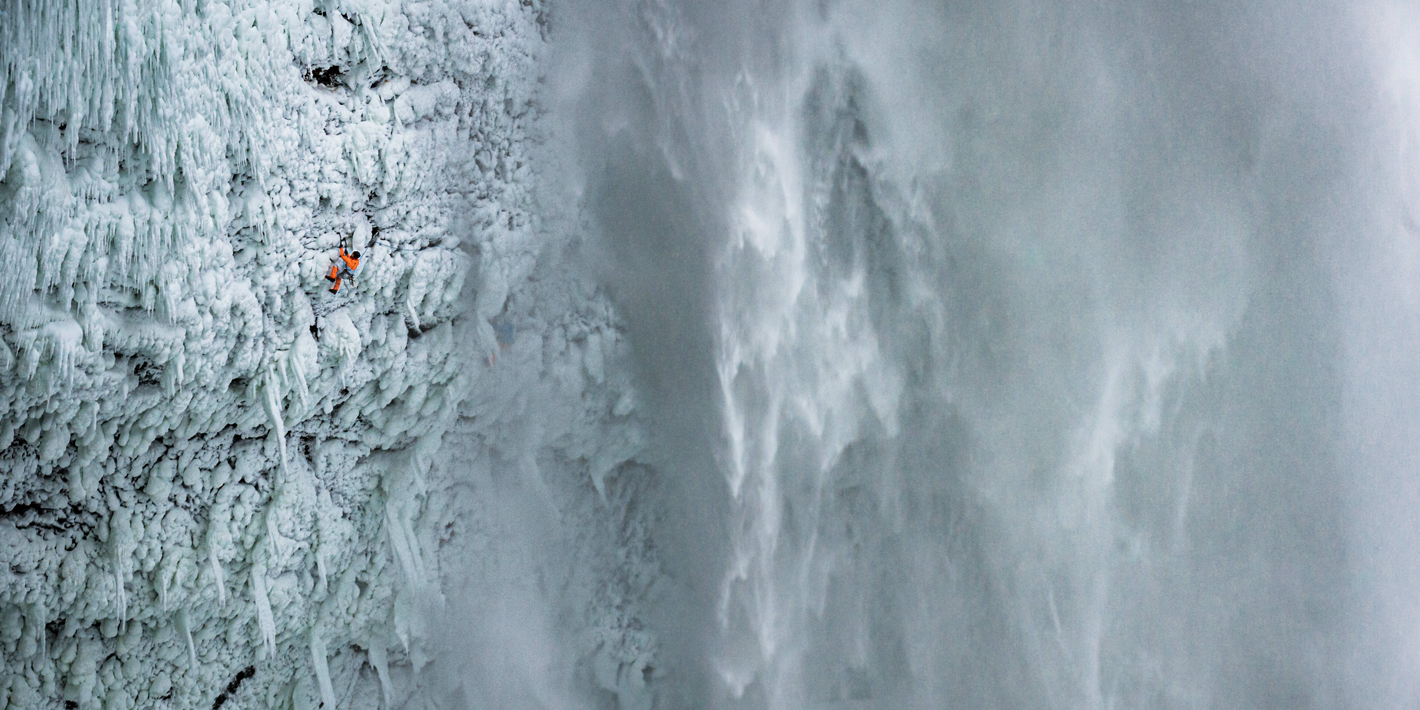 A climber, wearing Norrøna gear, scales an icy waterfall.  The water is frozen and the climber is small in the vast landscape. 
