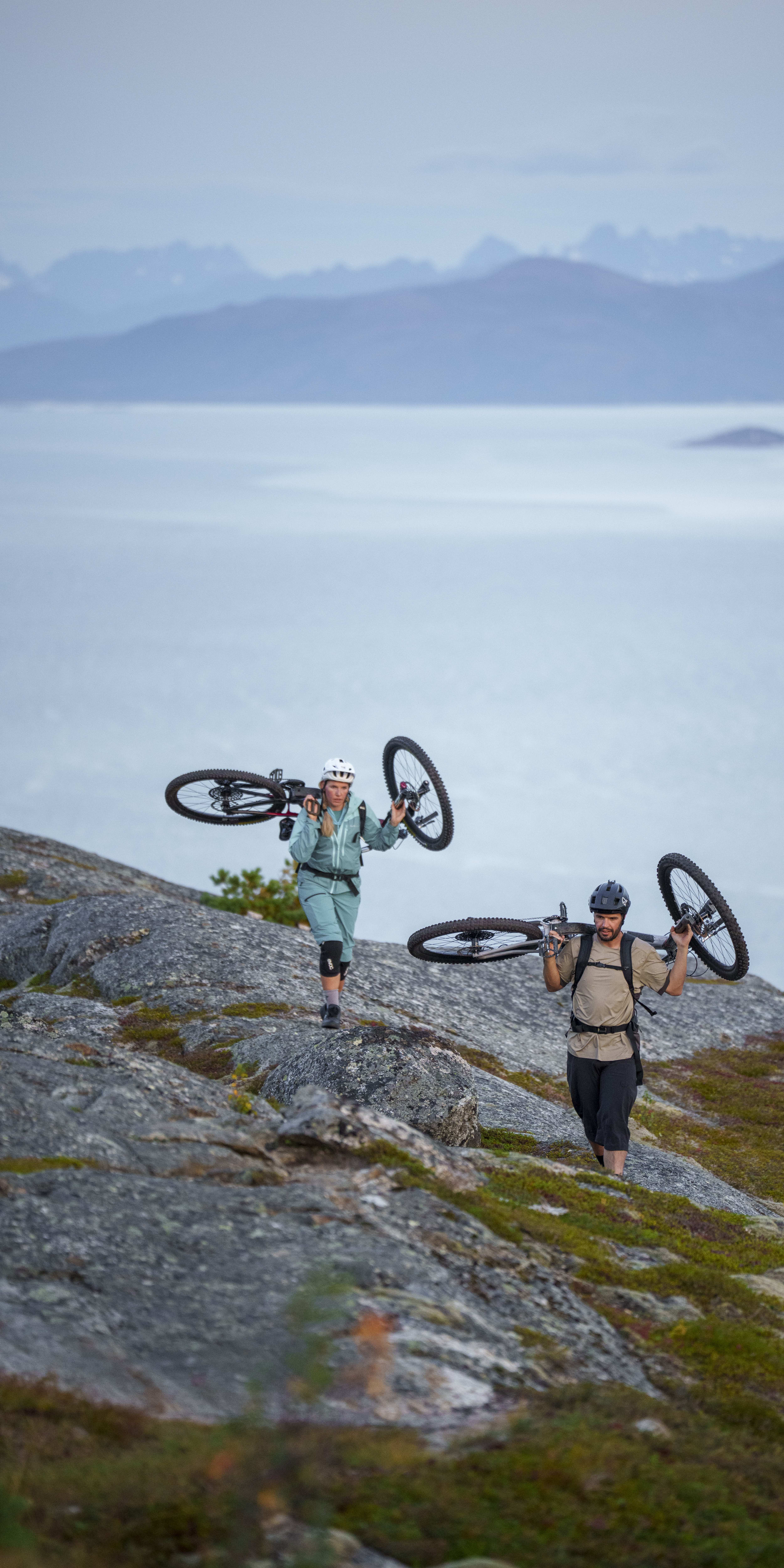A woman and a man walk uphill carrying mountain bikes. The woman wears a helmet and a light green Norrøna anorak and pants. Mountains and a body of water are in the background.
