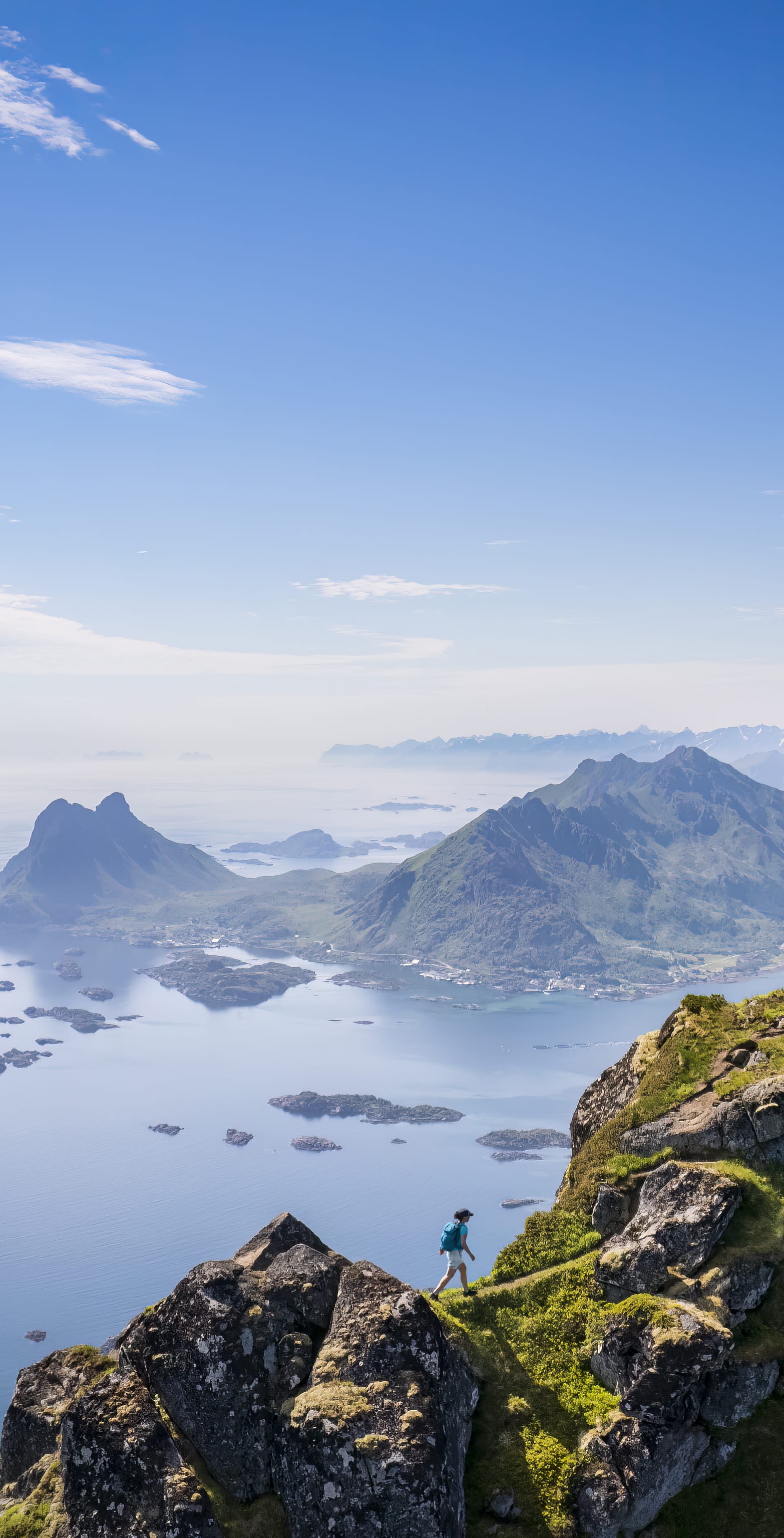 A person stands on a mountaintop overlooking a blue ocean with many rocky islands. The horizon is a hazy blue and the sky is clear and blue.