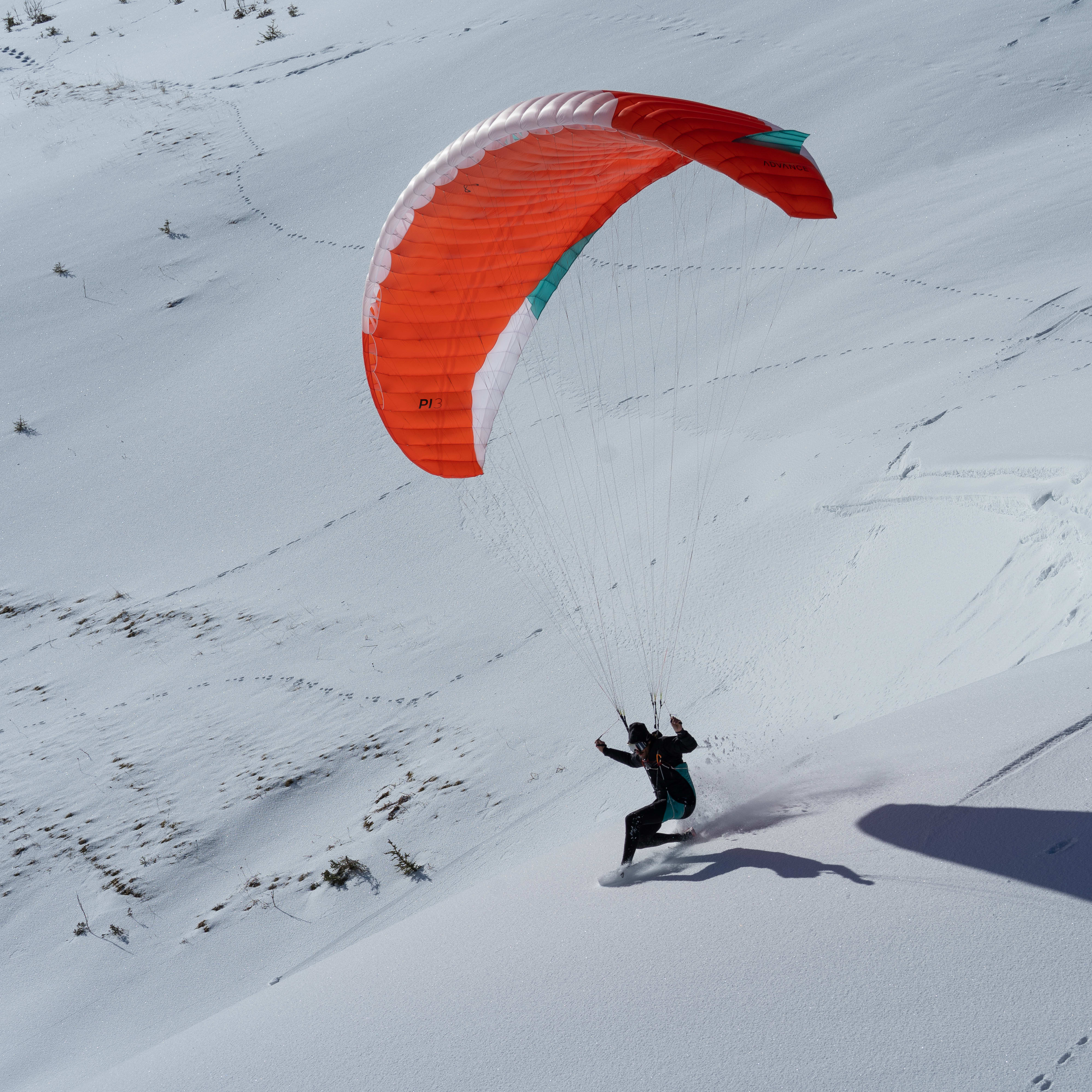 A person wearing a Norrøna jacket and a helmet is paragliding on a snowy mountain.  The parachute is red, white, and turquoise.