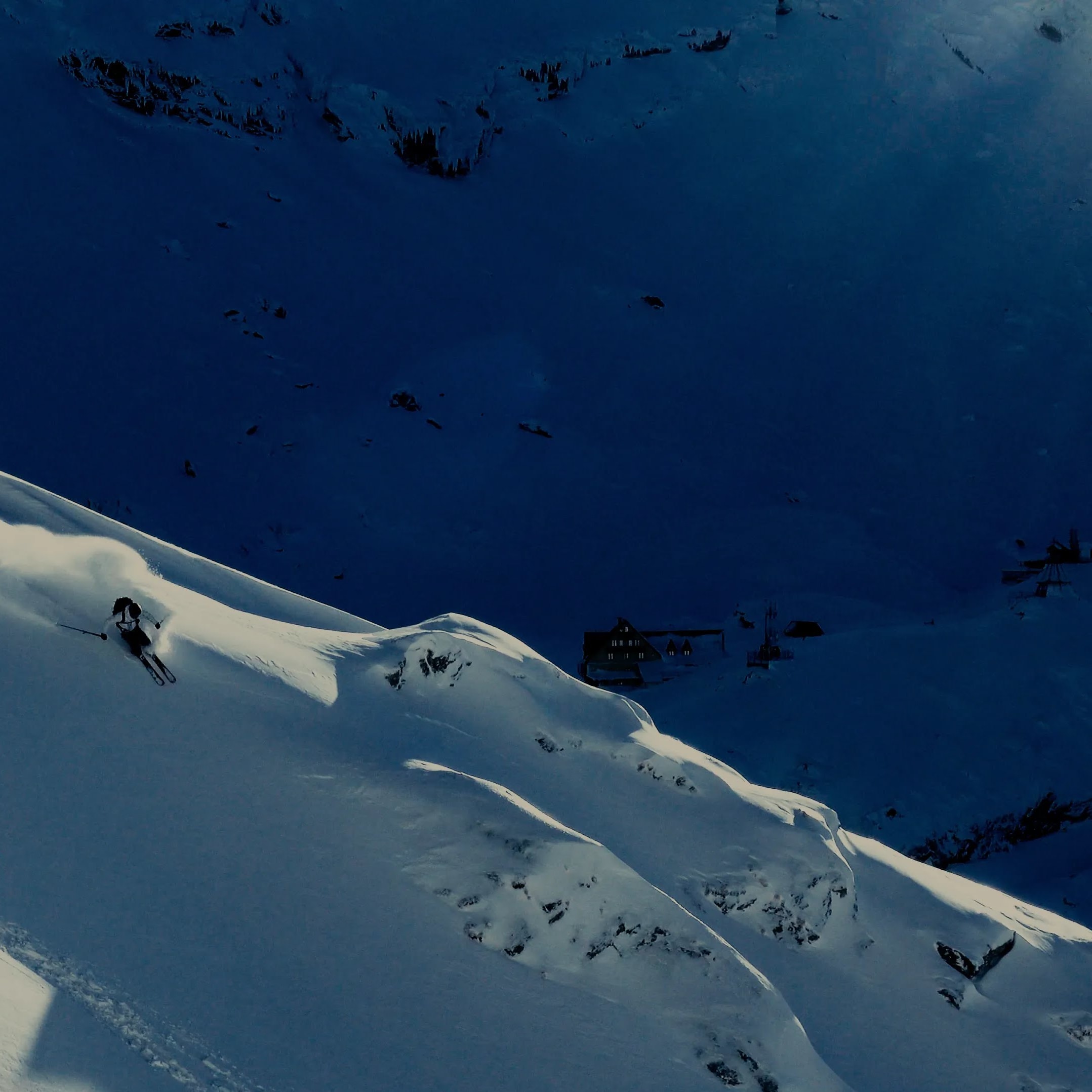 Skier in Norrøna gear carving down a steep, snow-covered mountain ridge, kicking up powder.