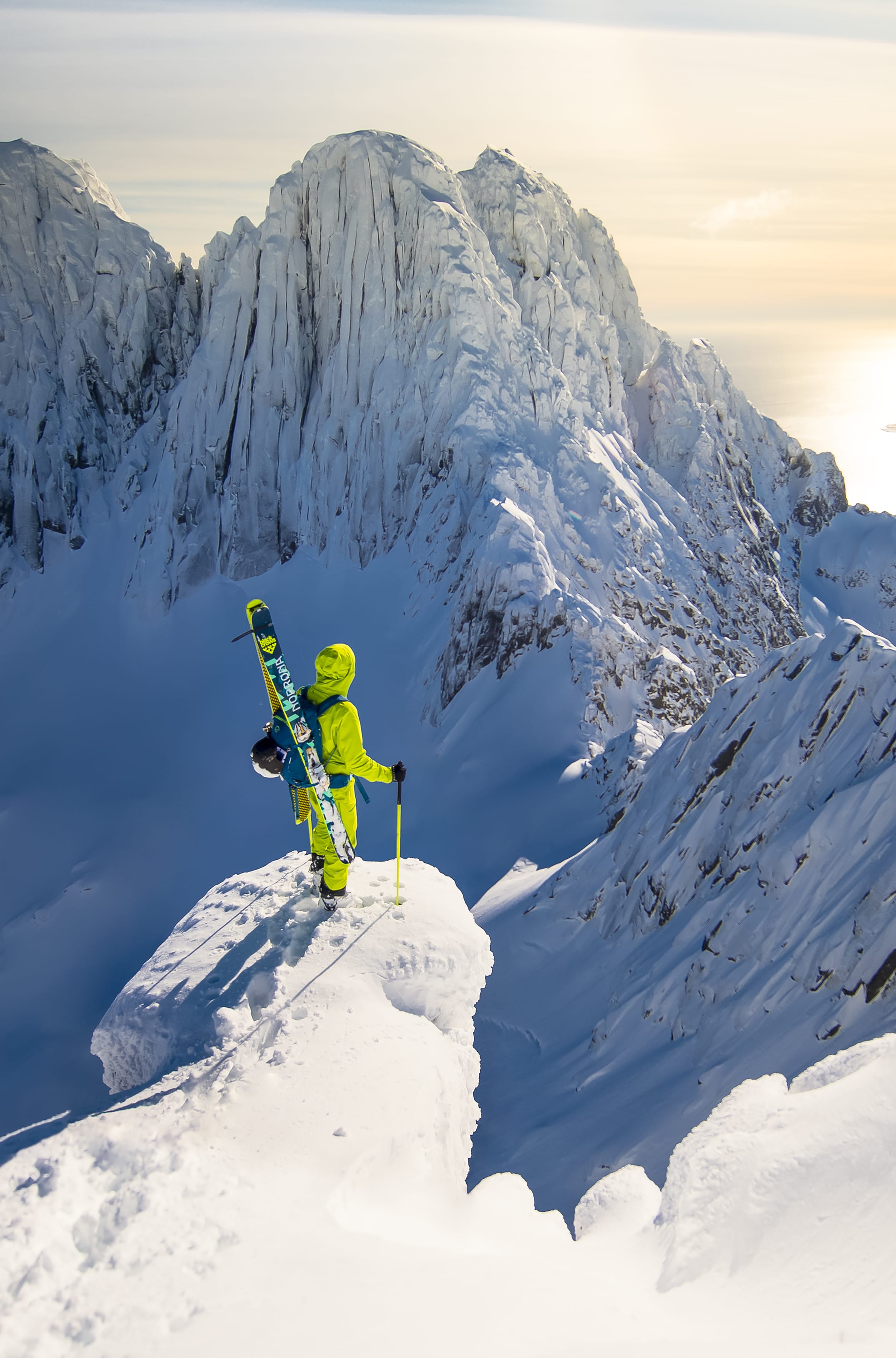 A person in yellow Norrøna gear stands on a snow-covered mountain peak with a pair of skis and ski poles. A vast expanse of snow-capped mountains and a glistening body of water stretch out below.