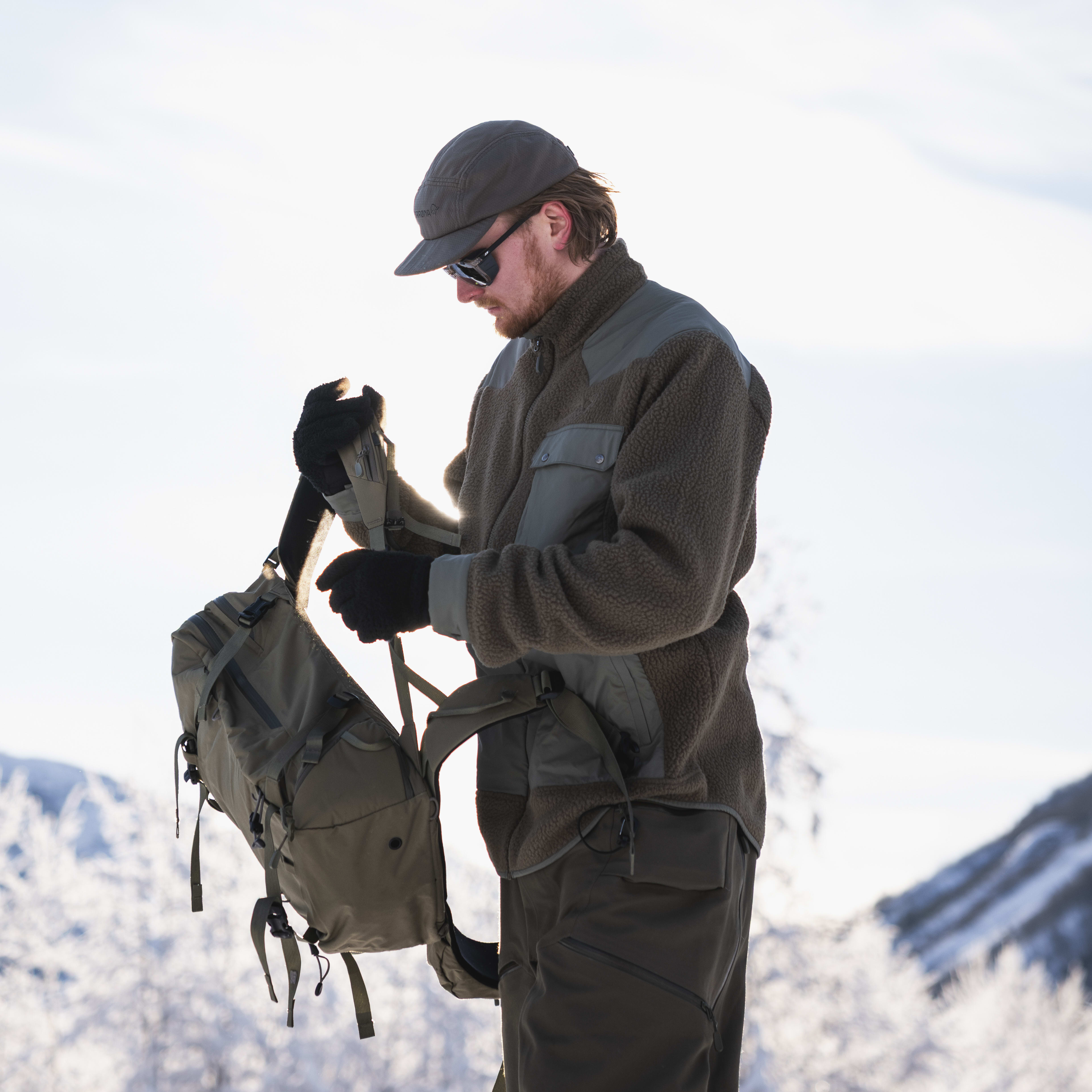 Man packing backpack in snowy landscape, wearing a hat, jacket, and pants, all from Norrøna.