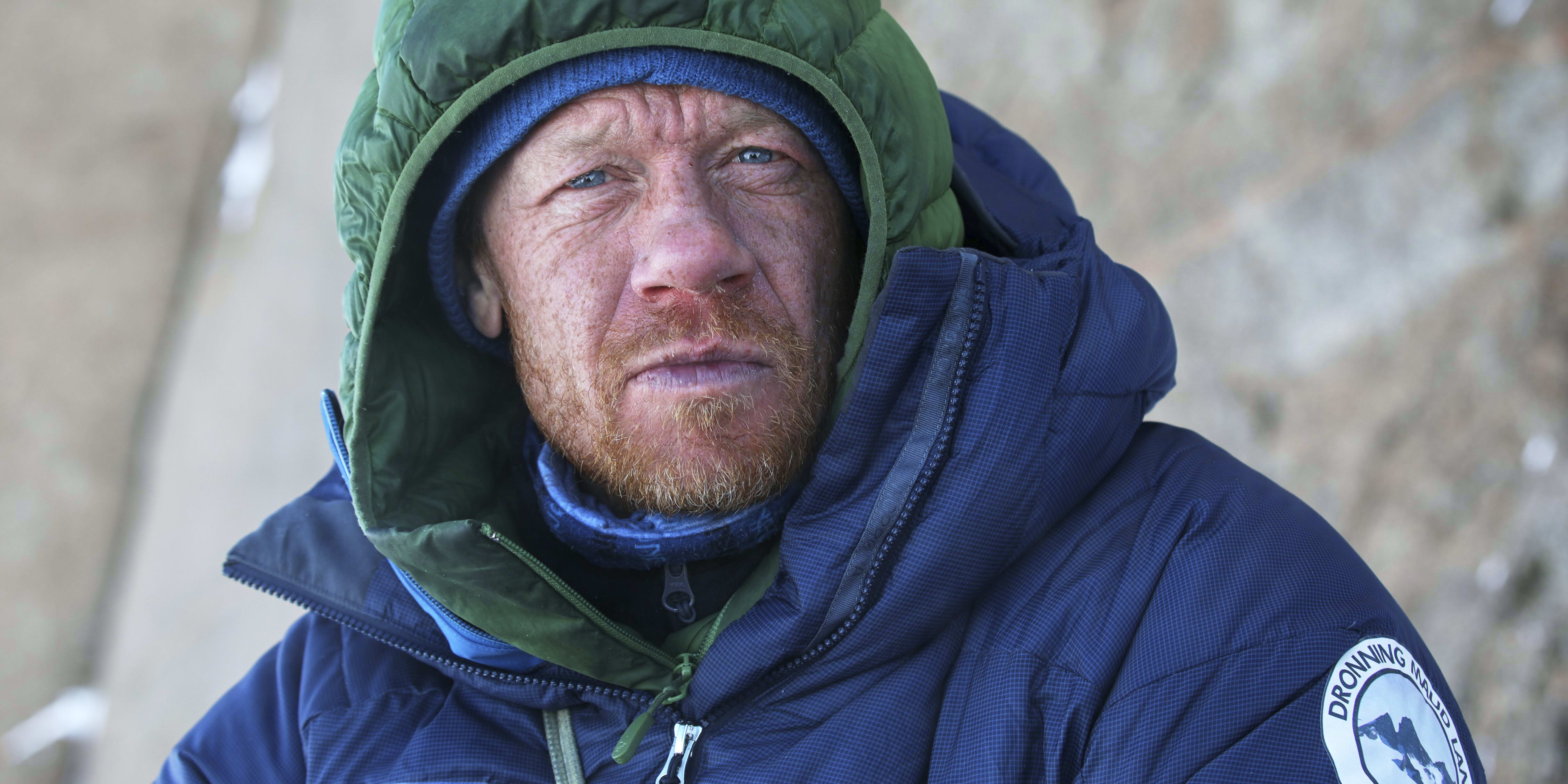 A man wearing a green and blue Norrøna jacket with a hood pulled up, looking to the right. He has a beard and blue eyes.