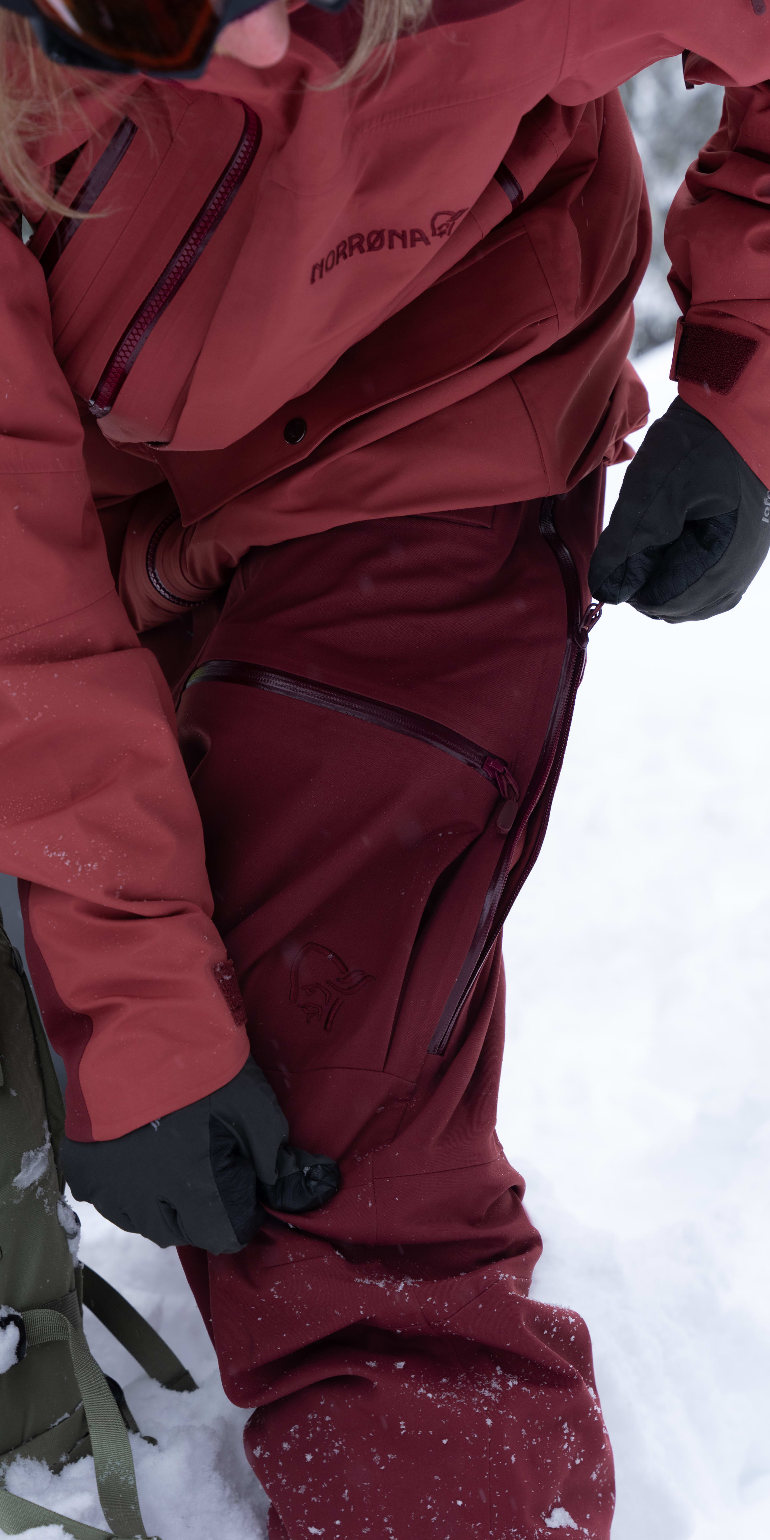 A person in the snow wearing a red Norrøna jacket and pants, zipping open a vent on the side of their pants.