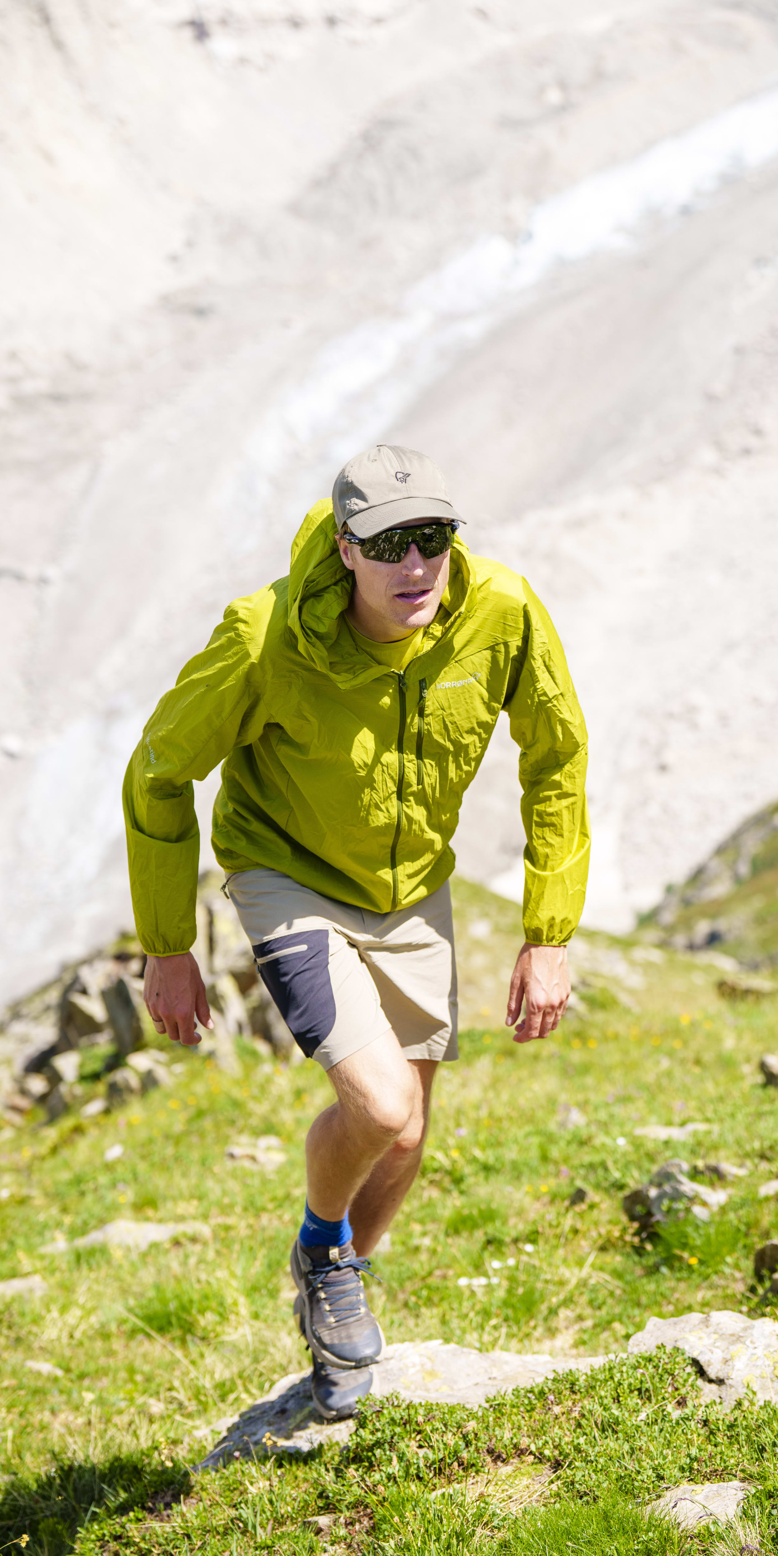 A man hikes uphill toward a glacier, wearing a green Norrøna jacket, tan Norrøna shorts, sunglasses, a cap, and trail shoes.

