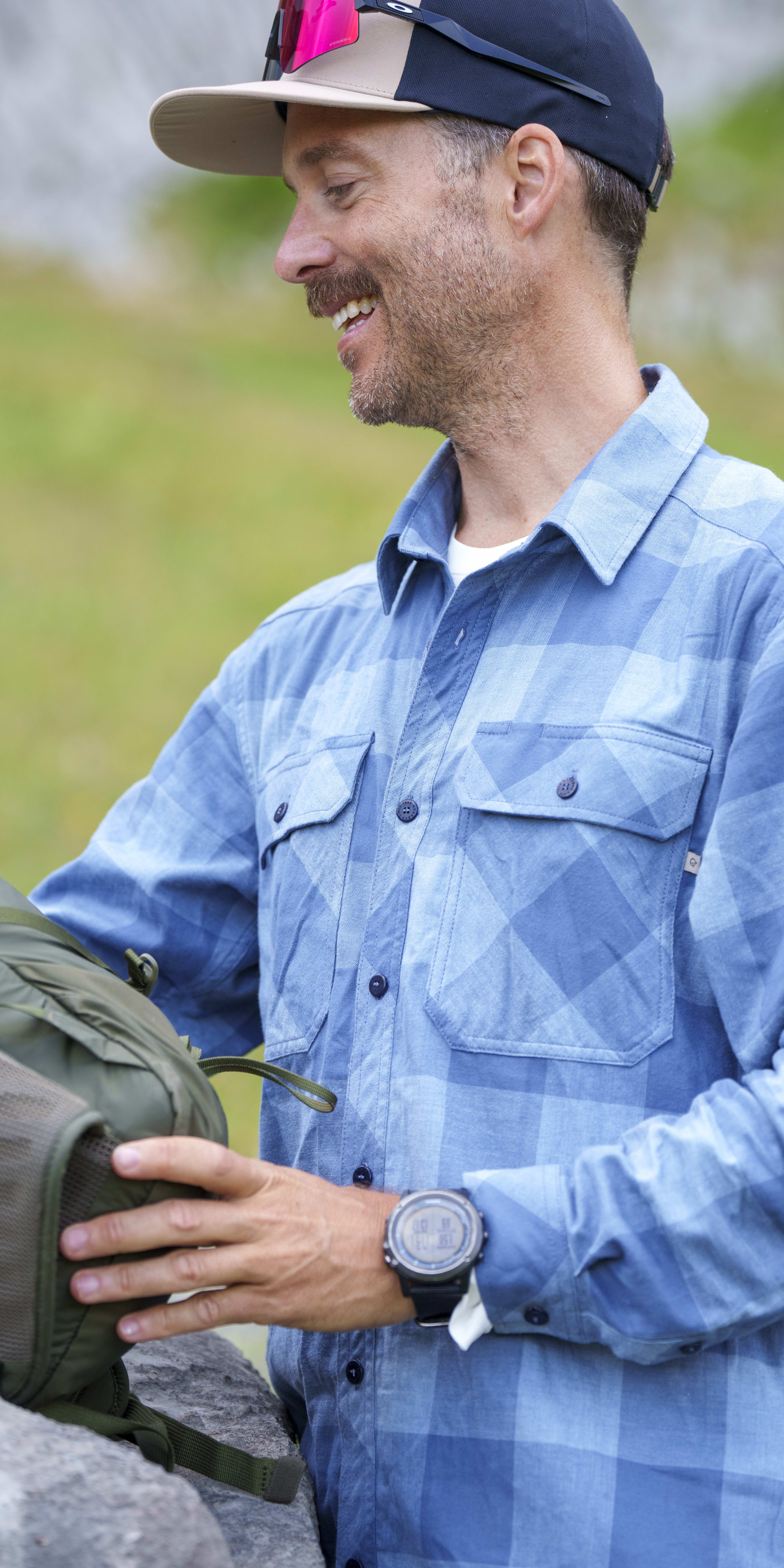 A smiling man with a beard and hat adjusts a green backpack. He wears a blue Norrøna flannel shirt and a watch.
