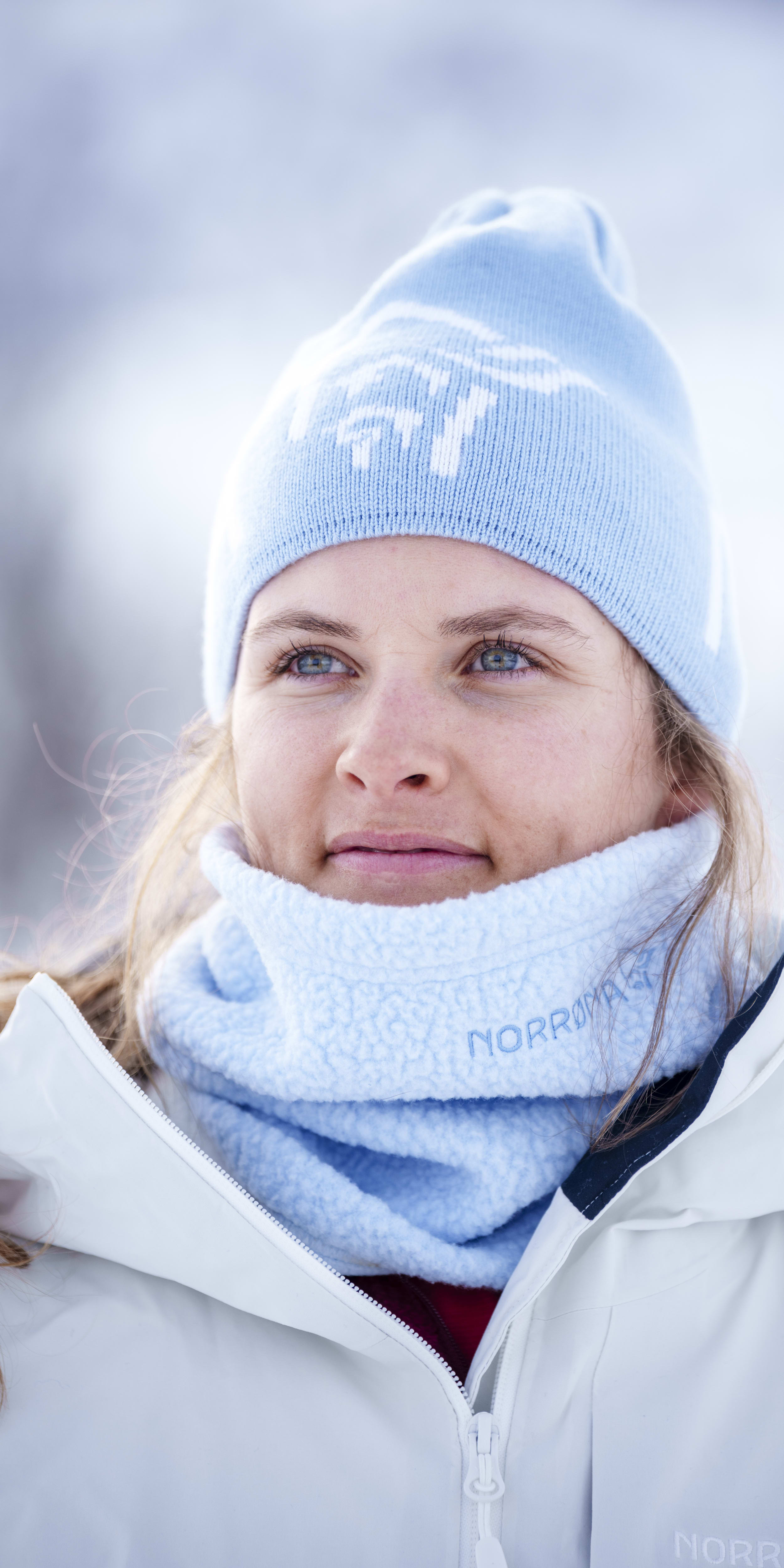 Portrait of a woman wearing a Norrøna beanie, neck warmer and white jacket against a snowy backdrop.