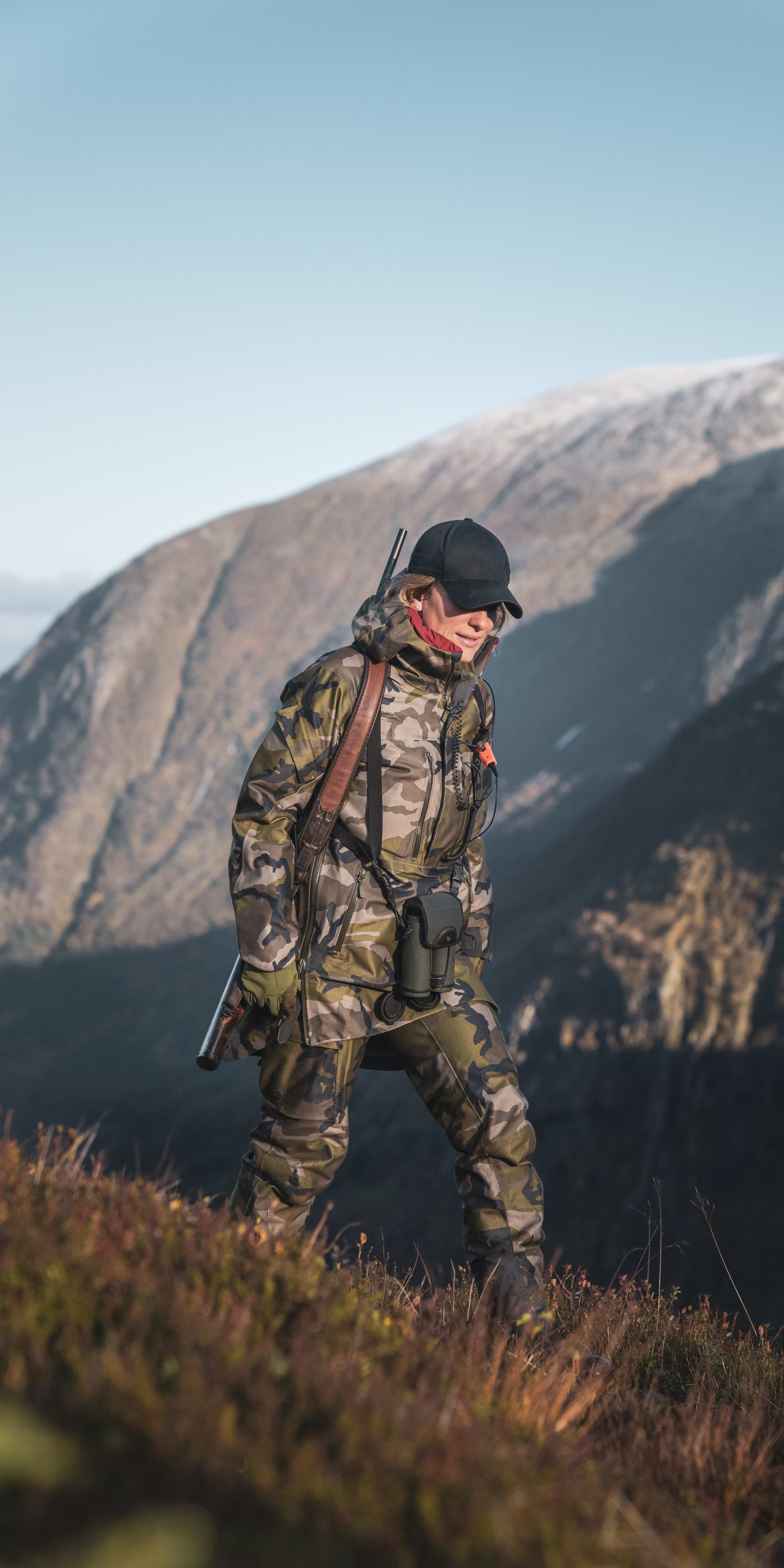 A person wearing camouflage Norrøna clothing and a hat walks through a grassy hillside with a rifle. 
