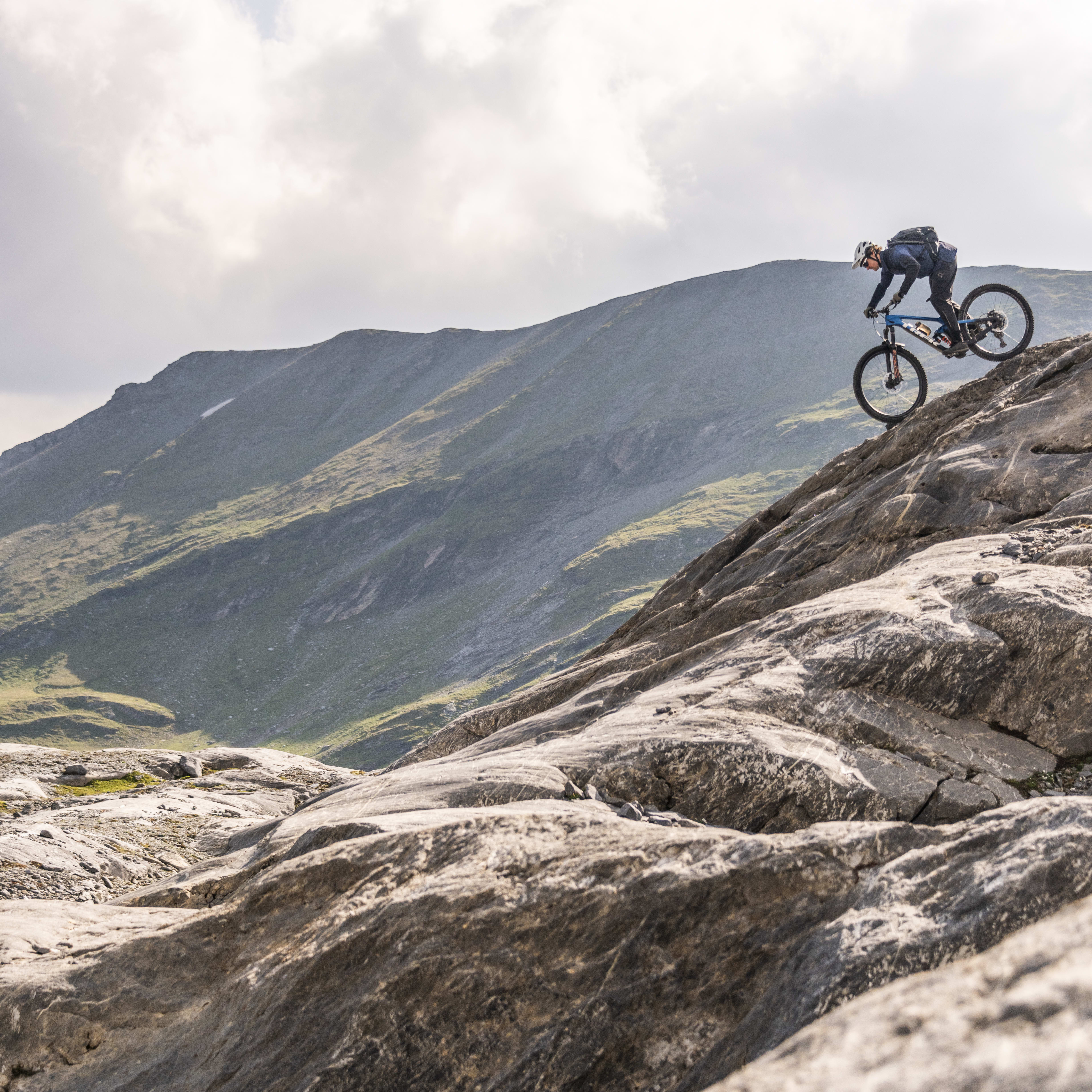 A person riding a mountain bike down a rocky mountain trail. The rider is wearing a helmet and a Norrøna backpack. The sky is cloudy.