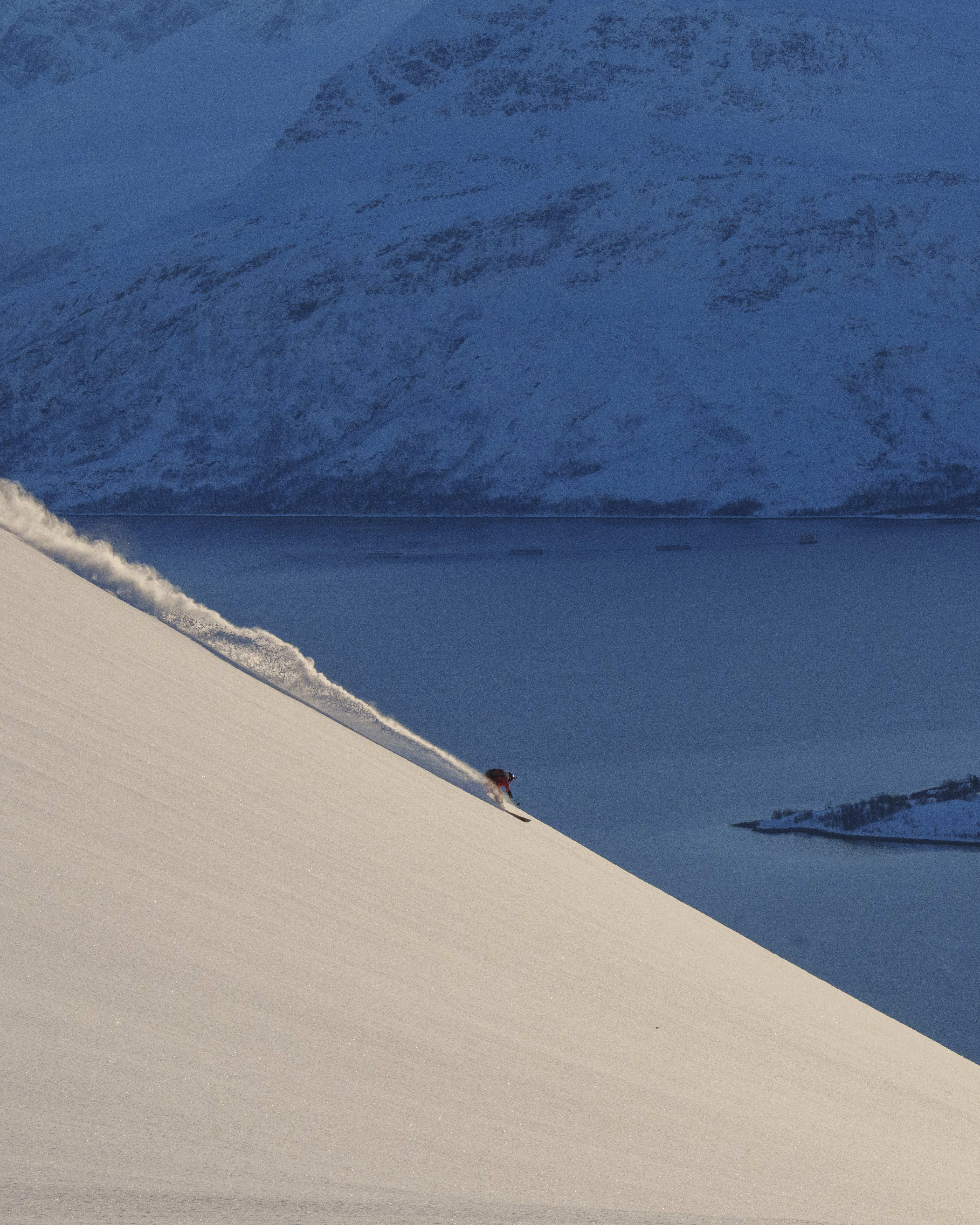 A skier in a Norrøna jacket descends a snowy slope, kicking up snow. A blue fjord and mountains are in the background.