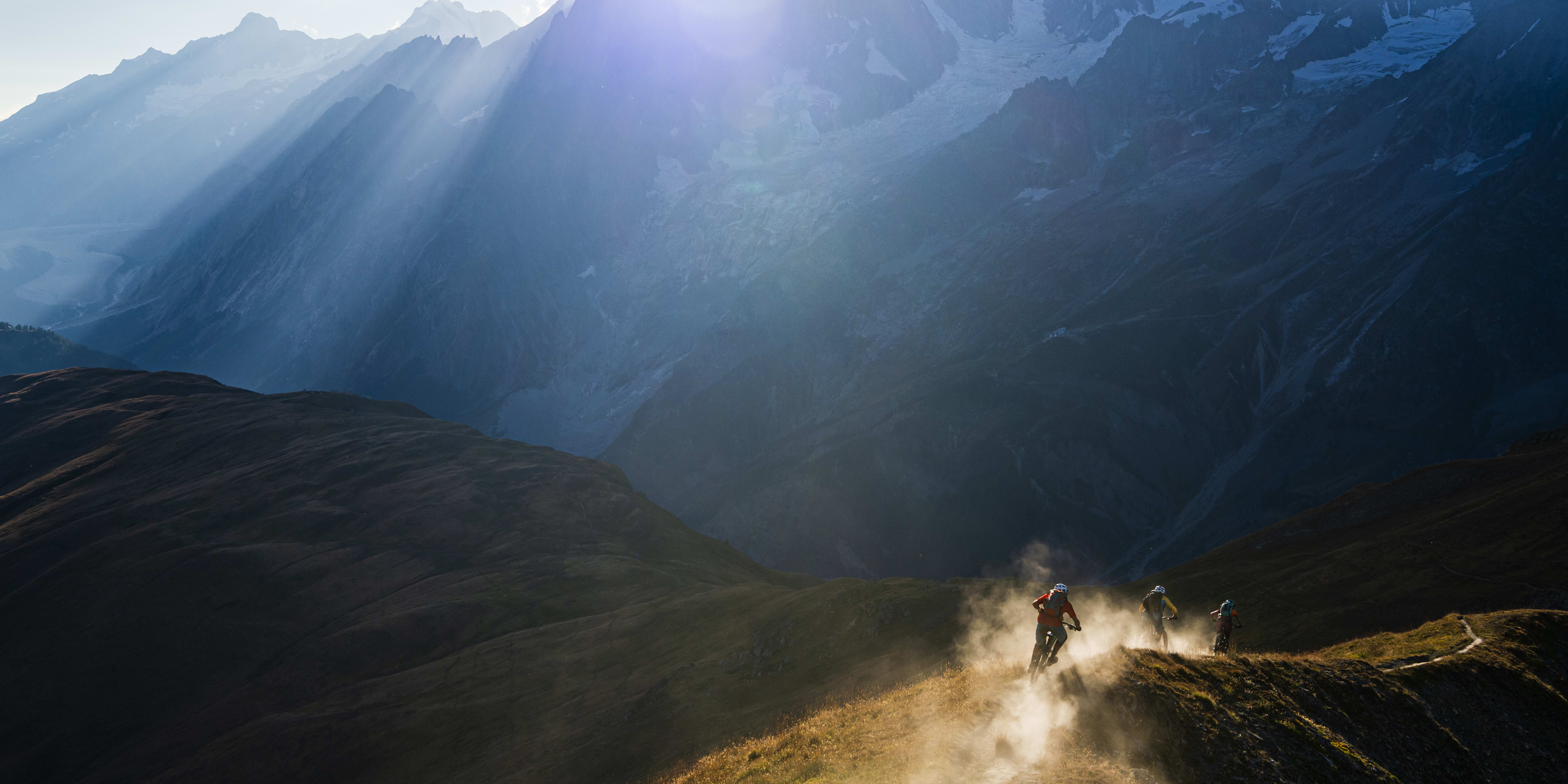 Three mountain bikers descending a trail in the French Alps. The sun shines through the clouds onto the snow-capped peaks. 
