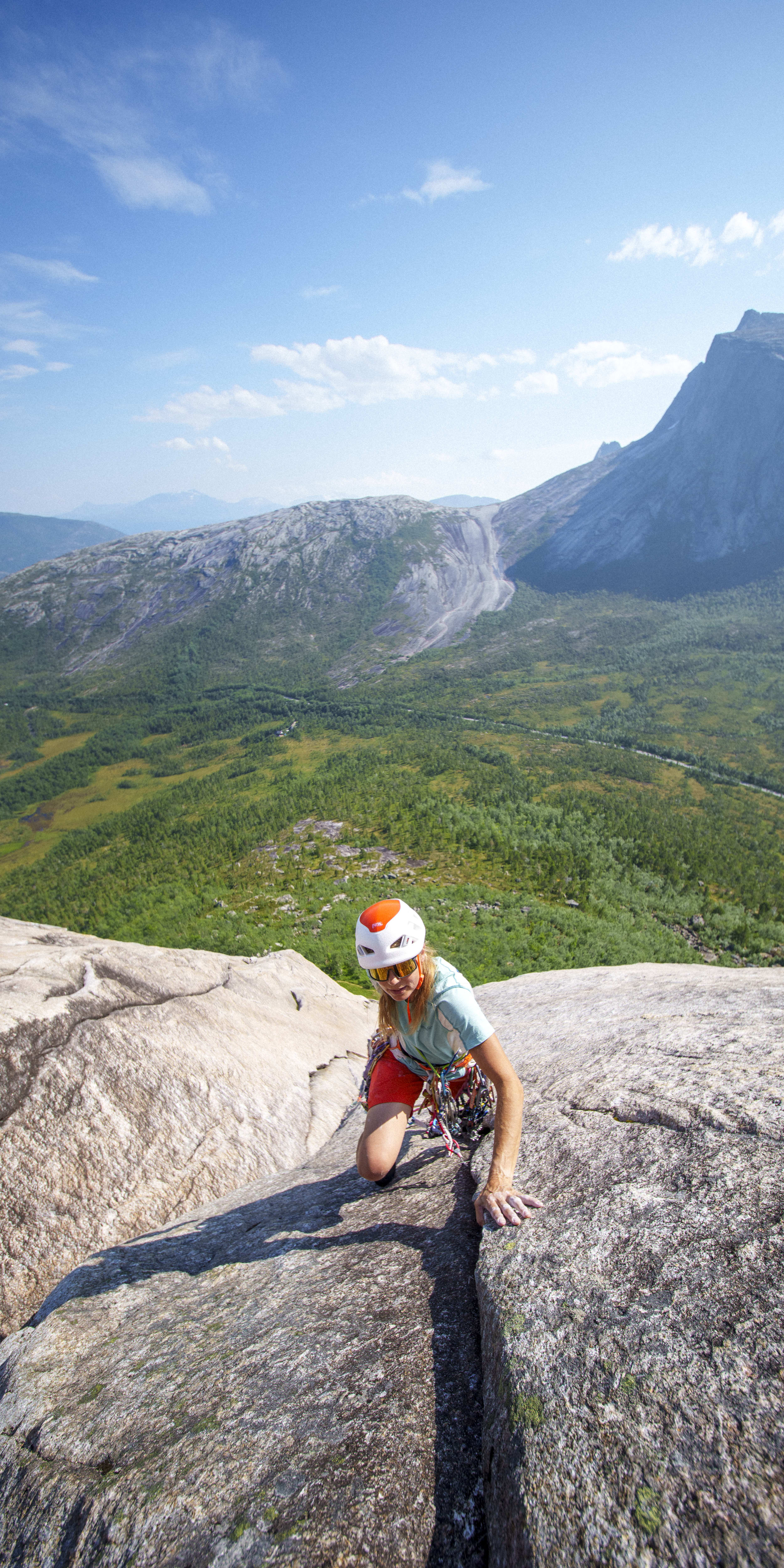 A climber with a helmet ascends a rock face. They wear Norrøna shorts and gear, with a valley and mountain visible in the background.
