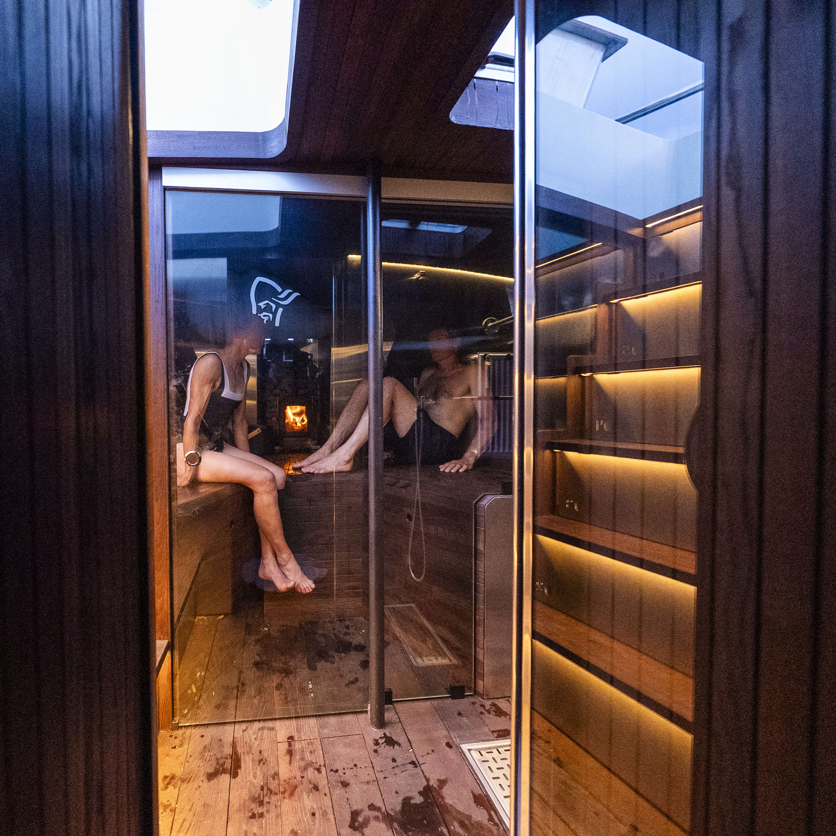 A glass-enclosed sauna featuring two people relaxing. One person wears a Norrøna swimsuit. Wooden interior and water droplets visible.