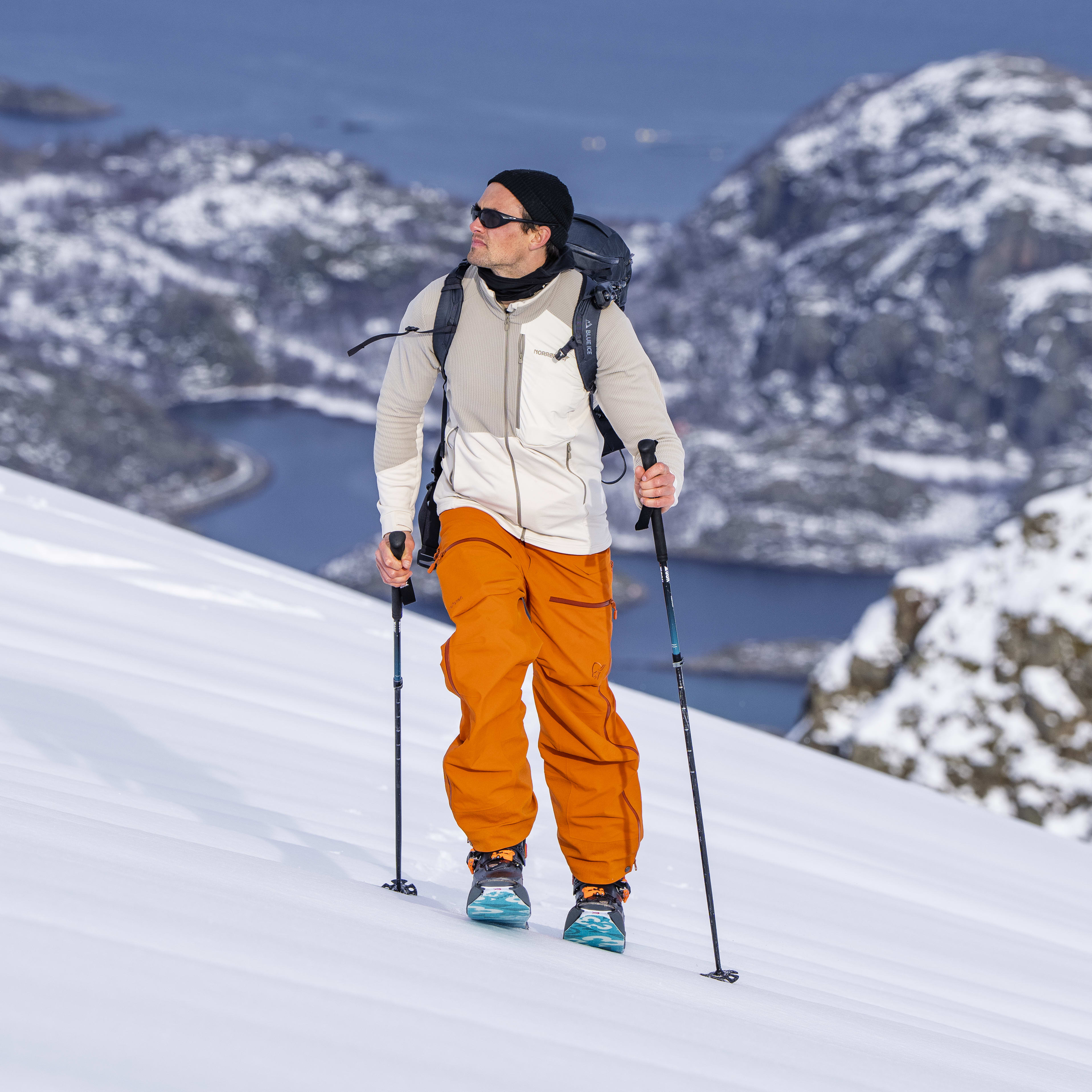 A man wearing Norrøna clothing and a backpack hikes through a snowy mountainous landscape.