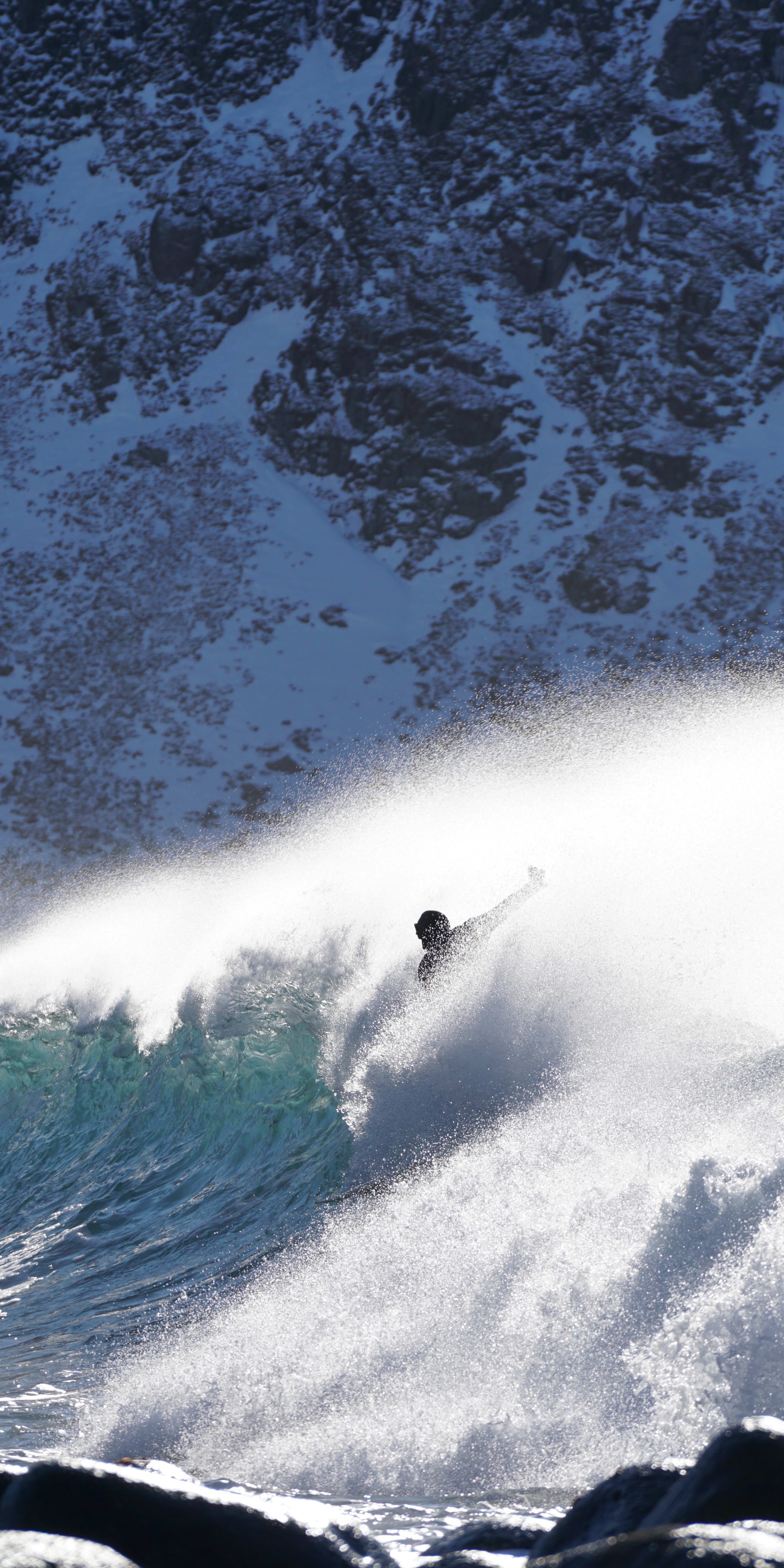 A person in a Norrøna jacket surfs a wave against a snowy mountain backdrop. 
