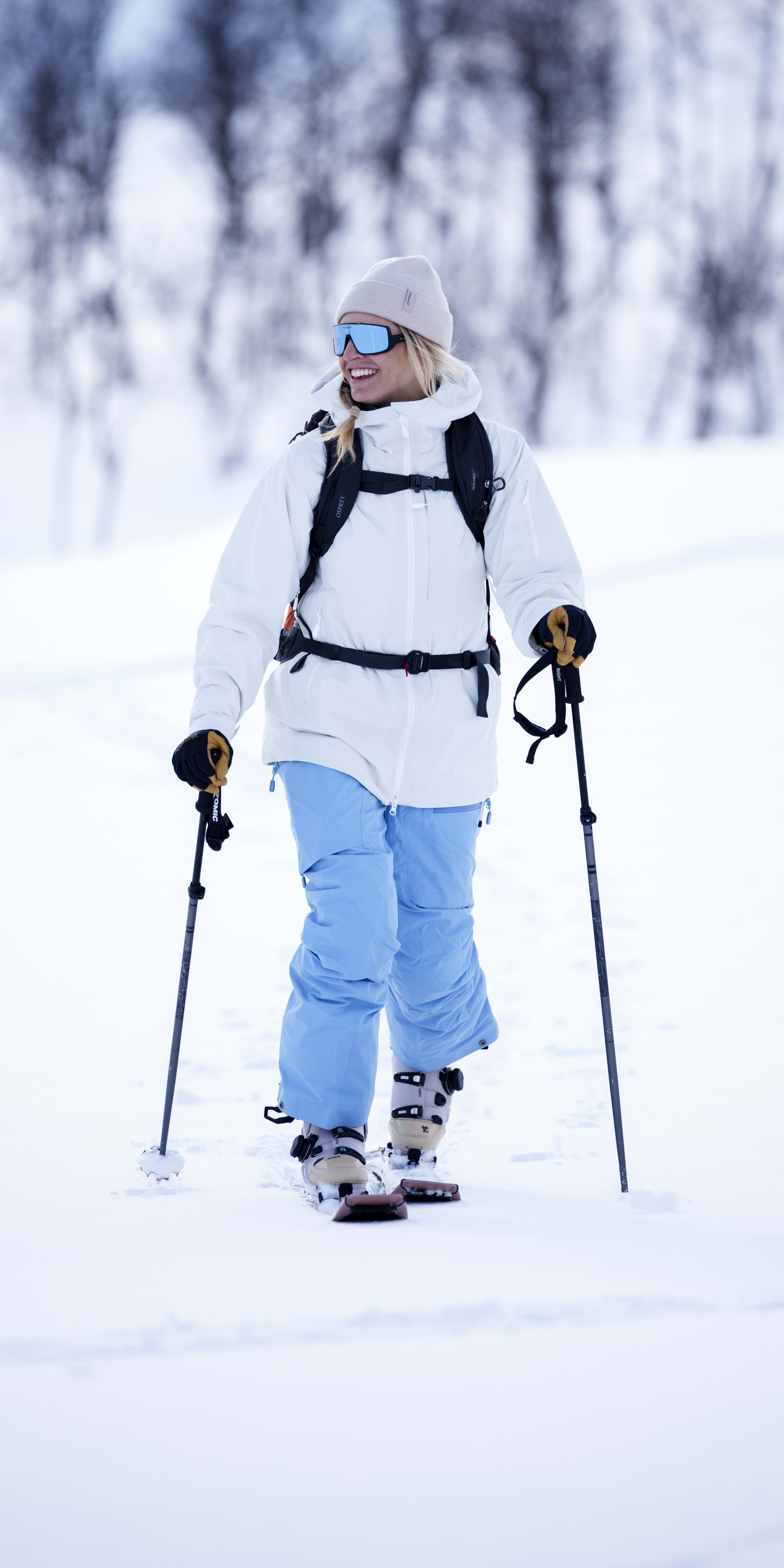 A smiling woman snowshoeing in a winter landscape. She's wearing a beanie, goggles, a Norrøna jacket and pants, and a backpack.
