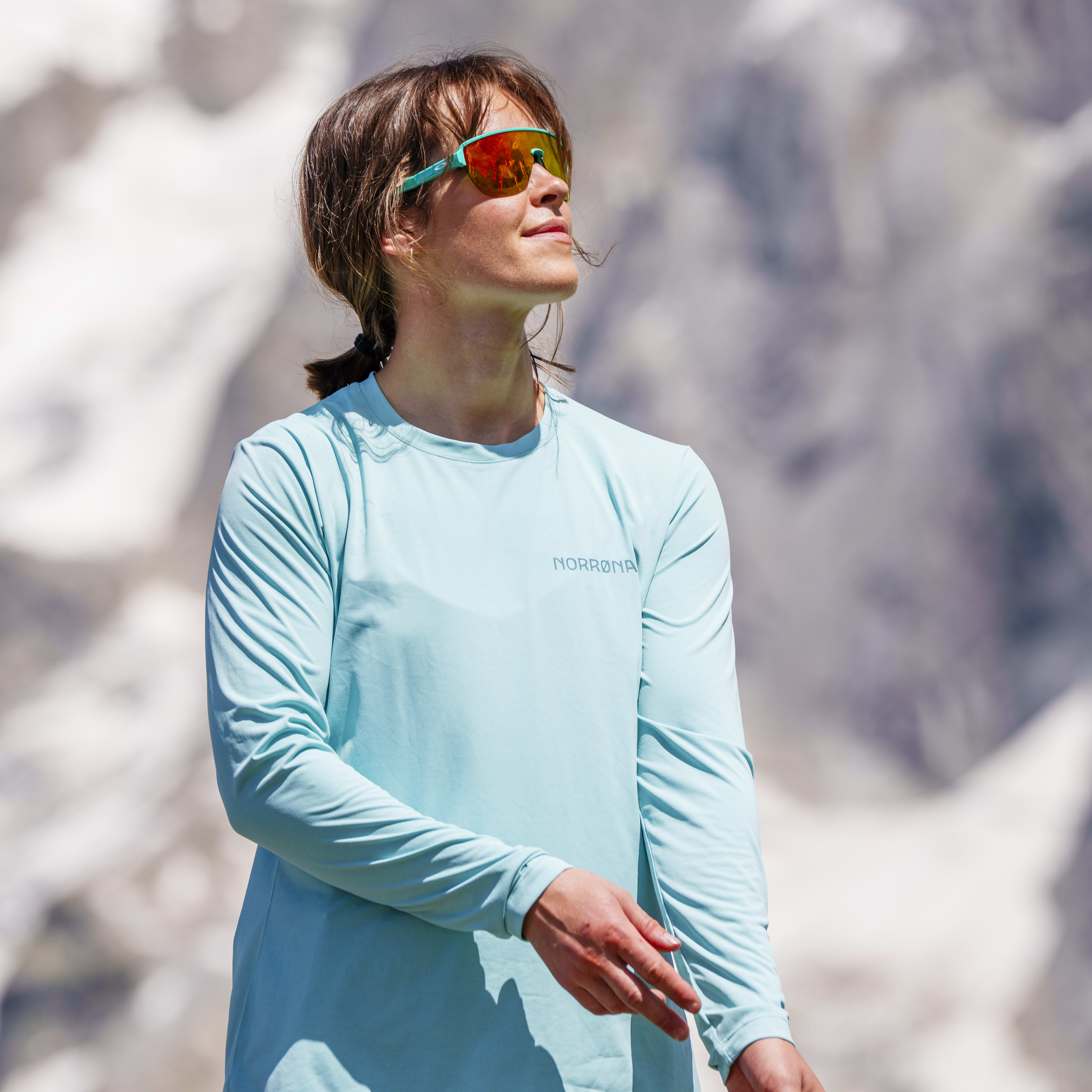 A woman in a turquoise Norrøna shirt and shorts, and sunglasses, gazes upwards, against a blurry mountain background.