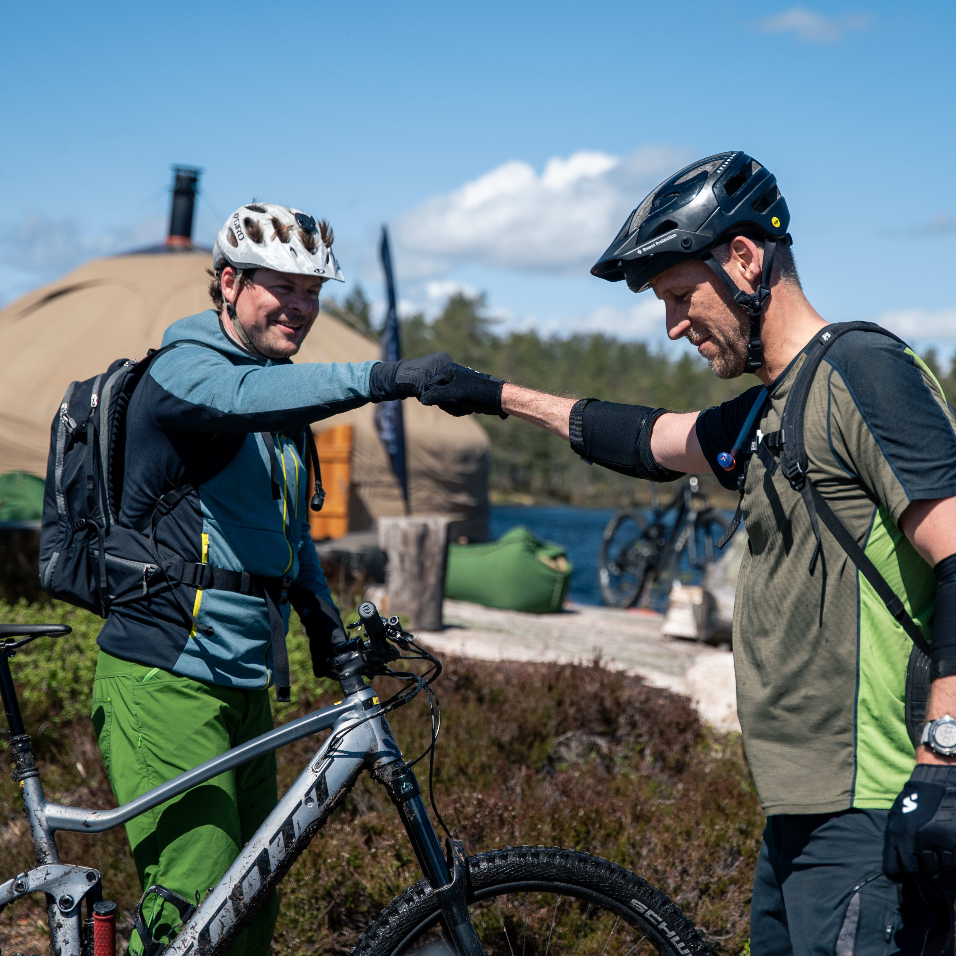 Two mountain bikers touch gloves in a fist bump on a trail. One wears a Norrøna hoodie and green pants. Bikes and a tent are visible.
