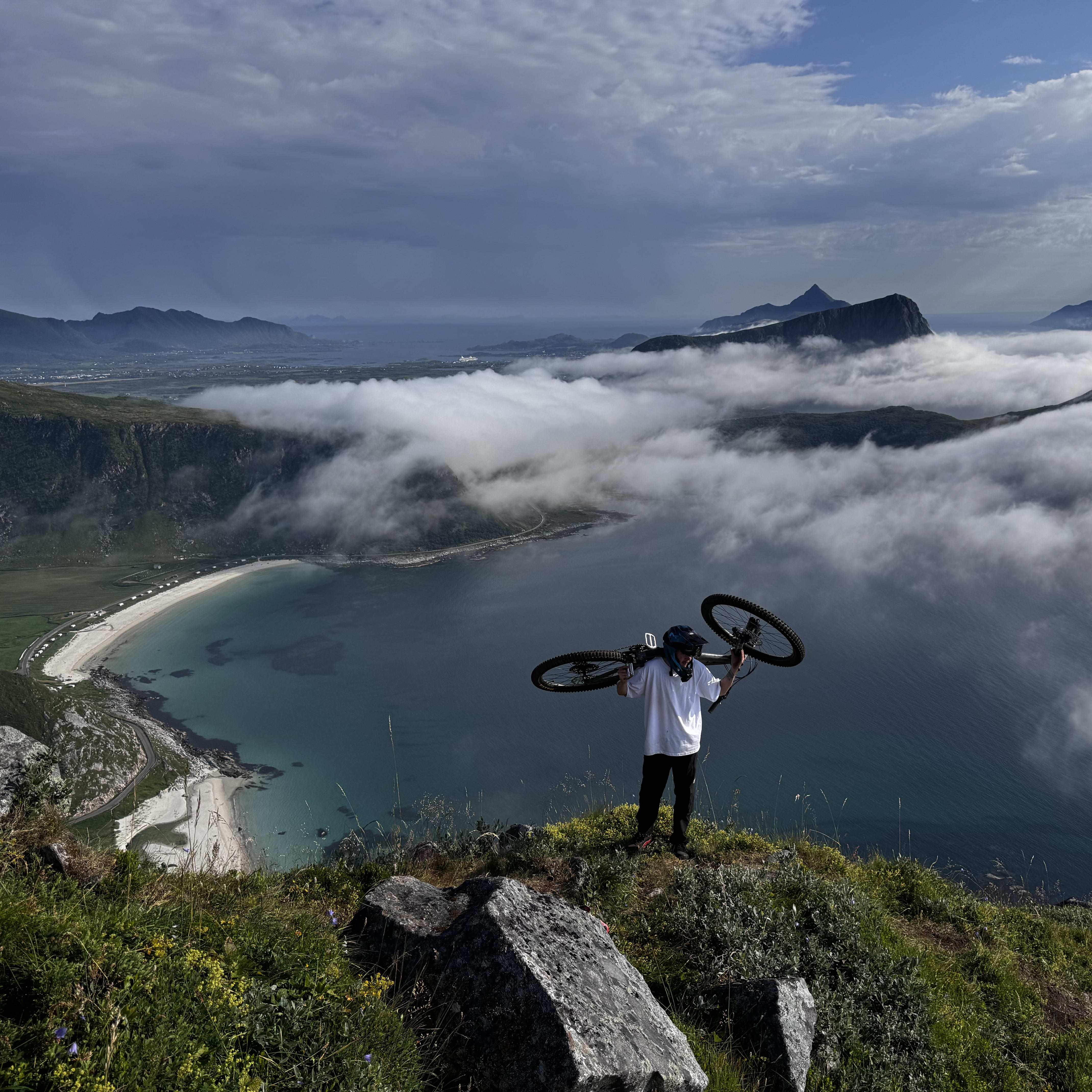 A person in a Norrøna shirt stands atop a mountain, carrying a bike, overlooking a coastline shrouded in clouds.