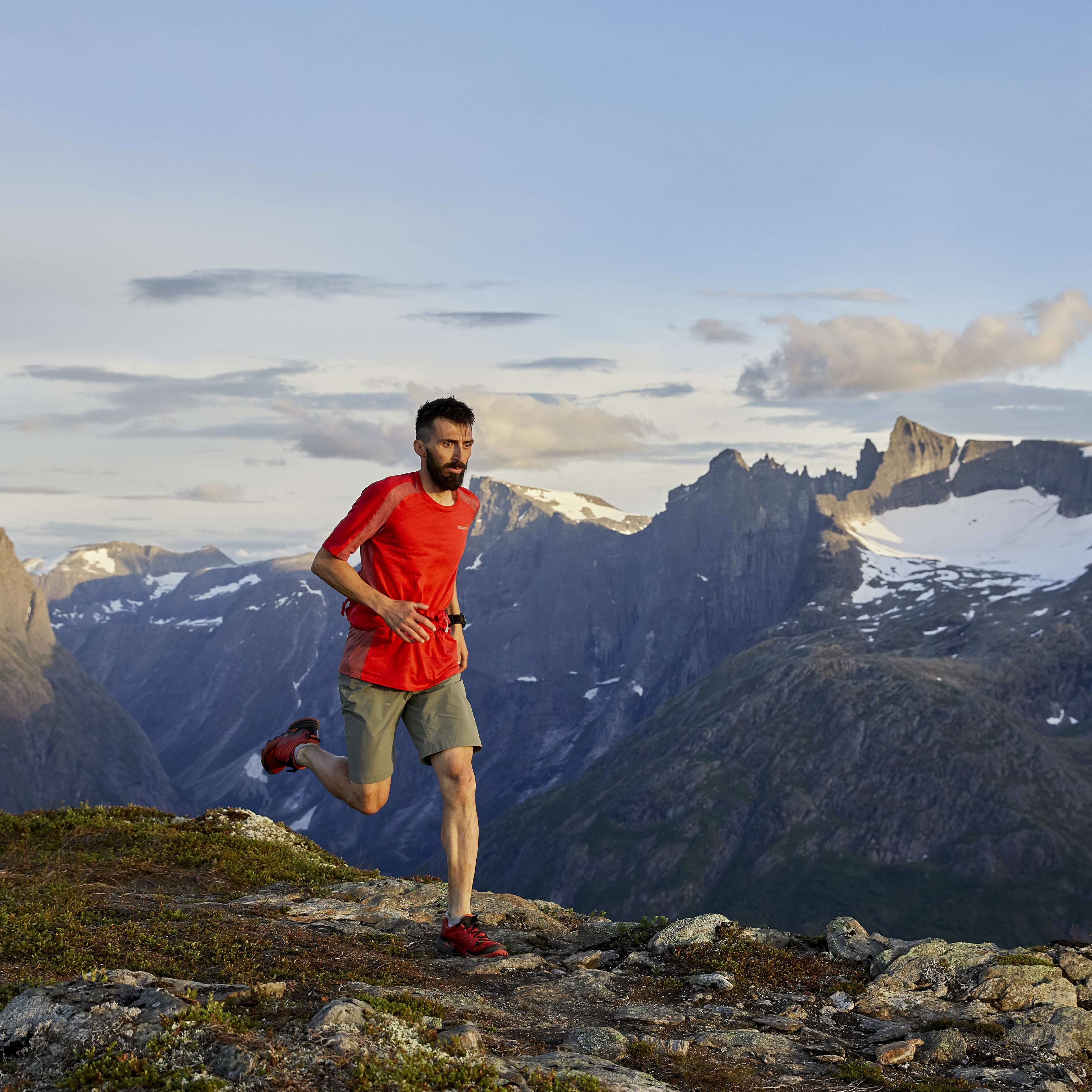 A man in red Norrøna gear runs on a mountain top, overlooking a vast mountainous landscape. 
