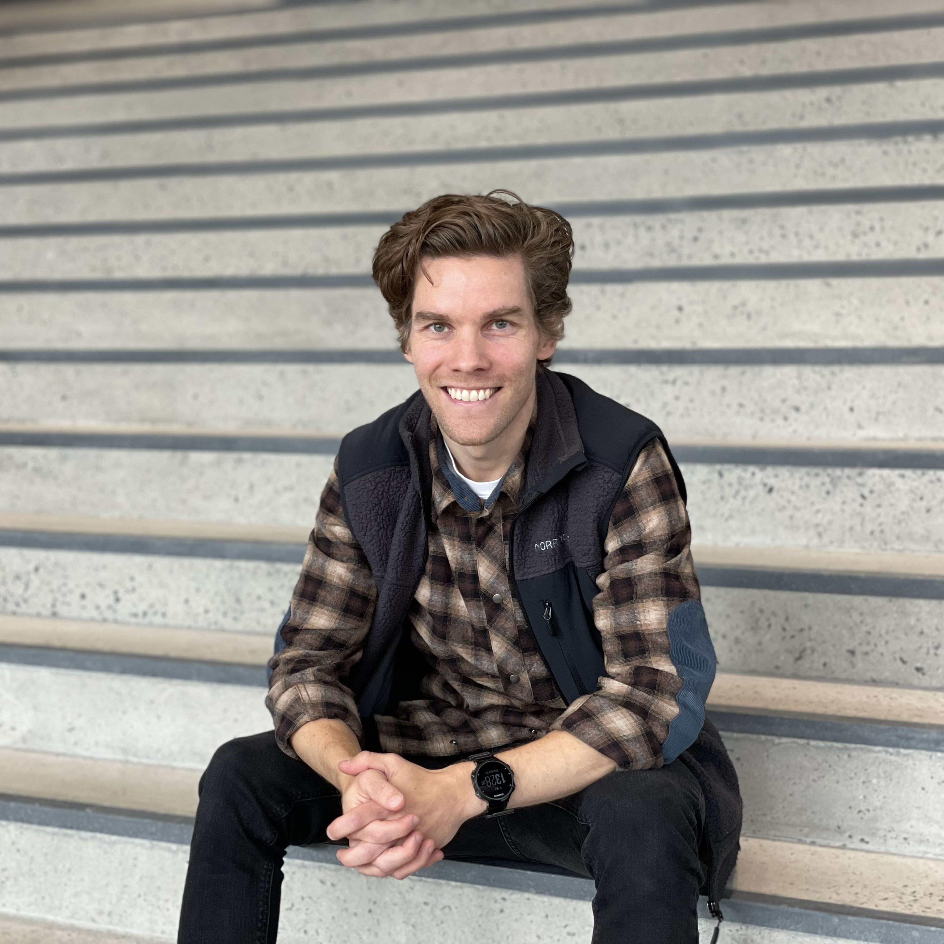 A man wearing a brown and black plaid shirt, a black fleece vest, and blue jeans sits on a set of concrete stairs with his hands clasped. He is smiling at the camera. His watch is visible on his left wrist.