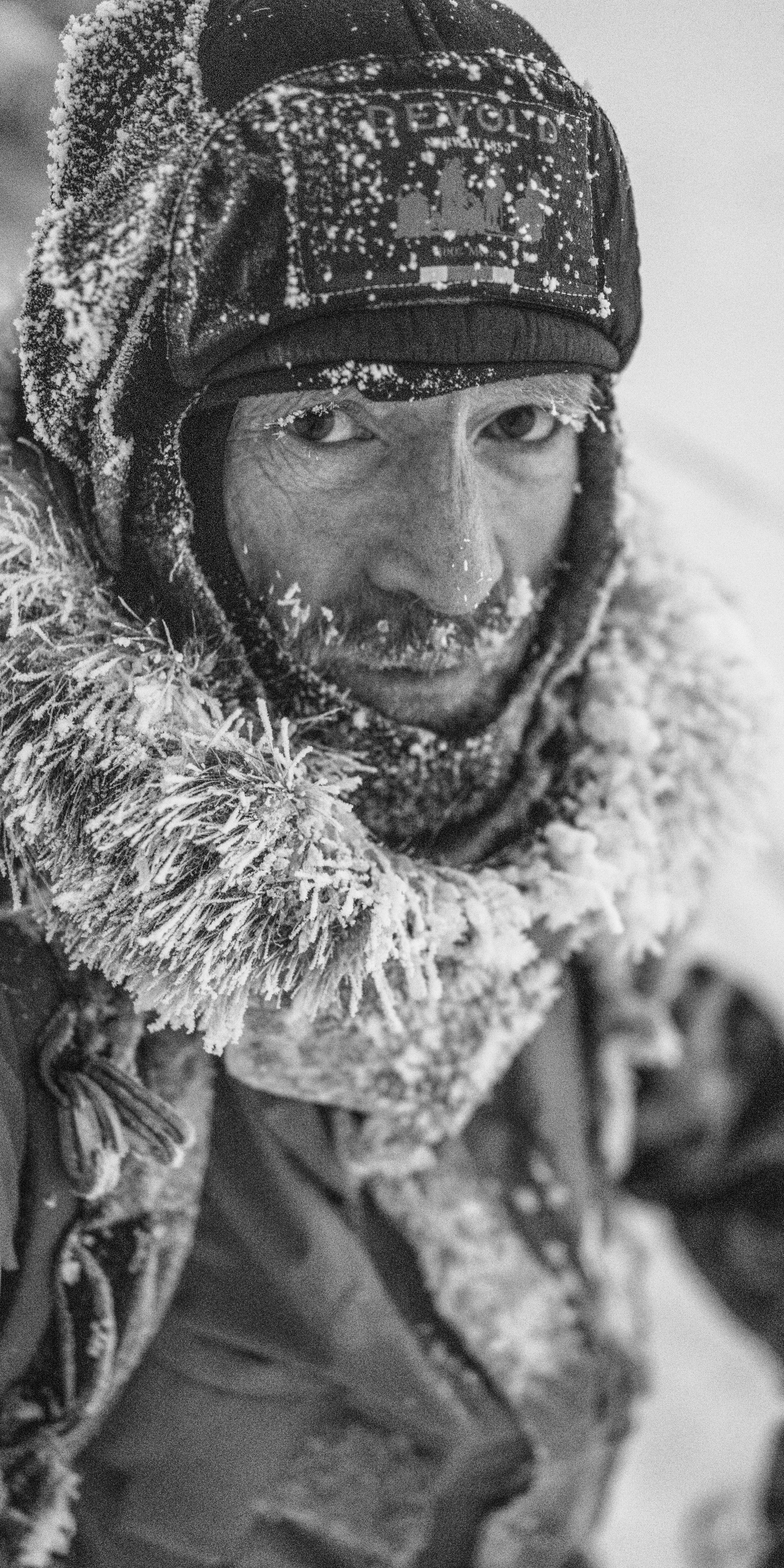 A man wearing a Norrøna hat and jacket is covered in snow and ice.  The cold weather creates a stark and textured image.