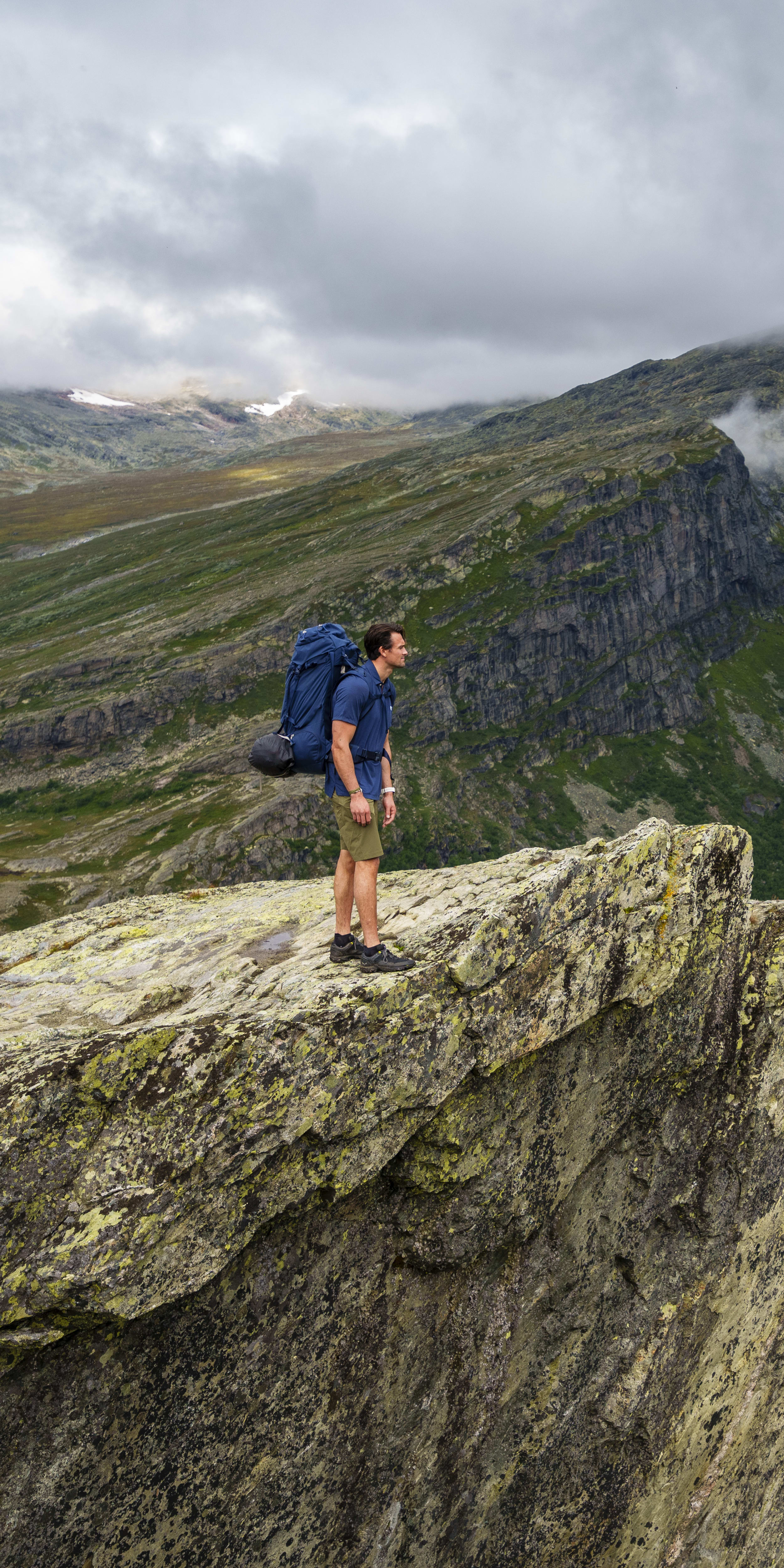 A man with a backpack stands on a rocky cliff edge overlooking a valley. The valley is covered in green vegetation and a winding road.  The mountain tops in the distance are covered in clouds.